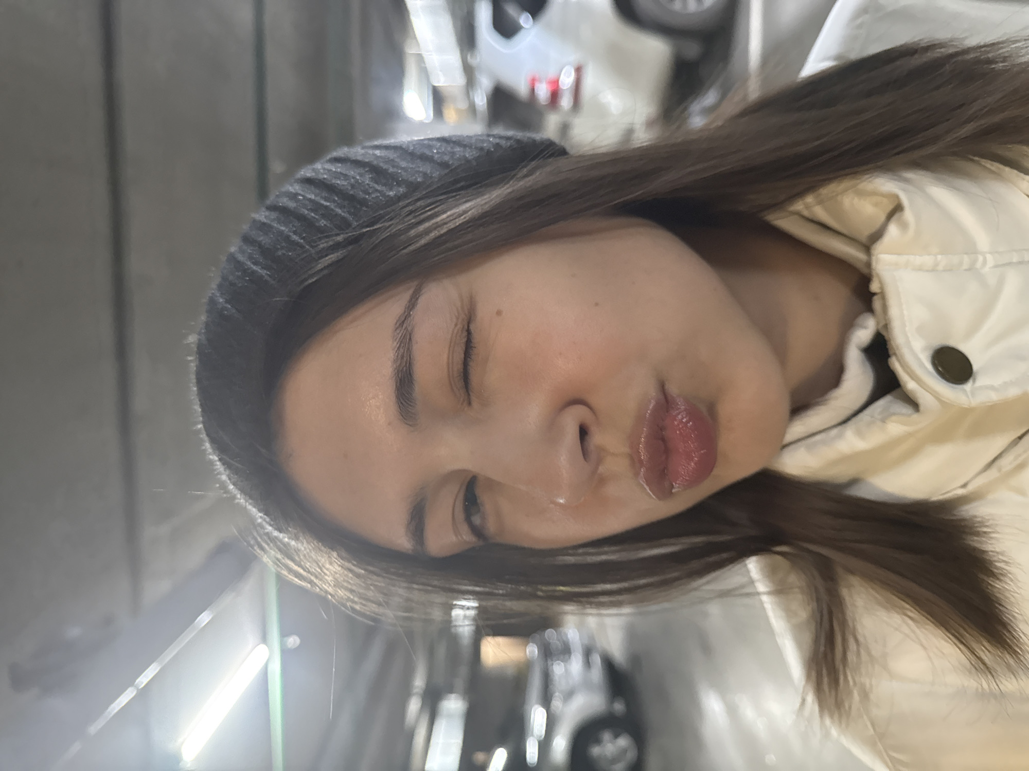 A woman with brown hair and a gray beanie is puckering her lips. She is wearing a light-colored jacket. The background appears to be a parking garage, with a car visible and fluorescent lighting. The exact location within Krasnoyarsk, Russia is unknown from the image alone.
