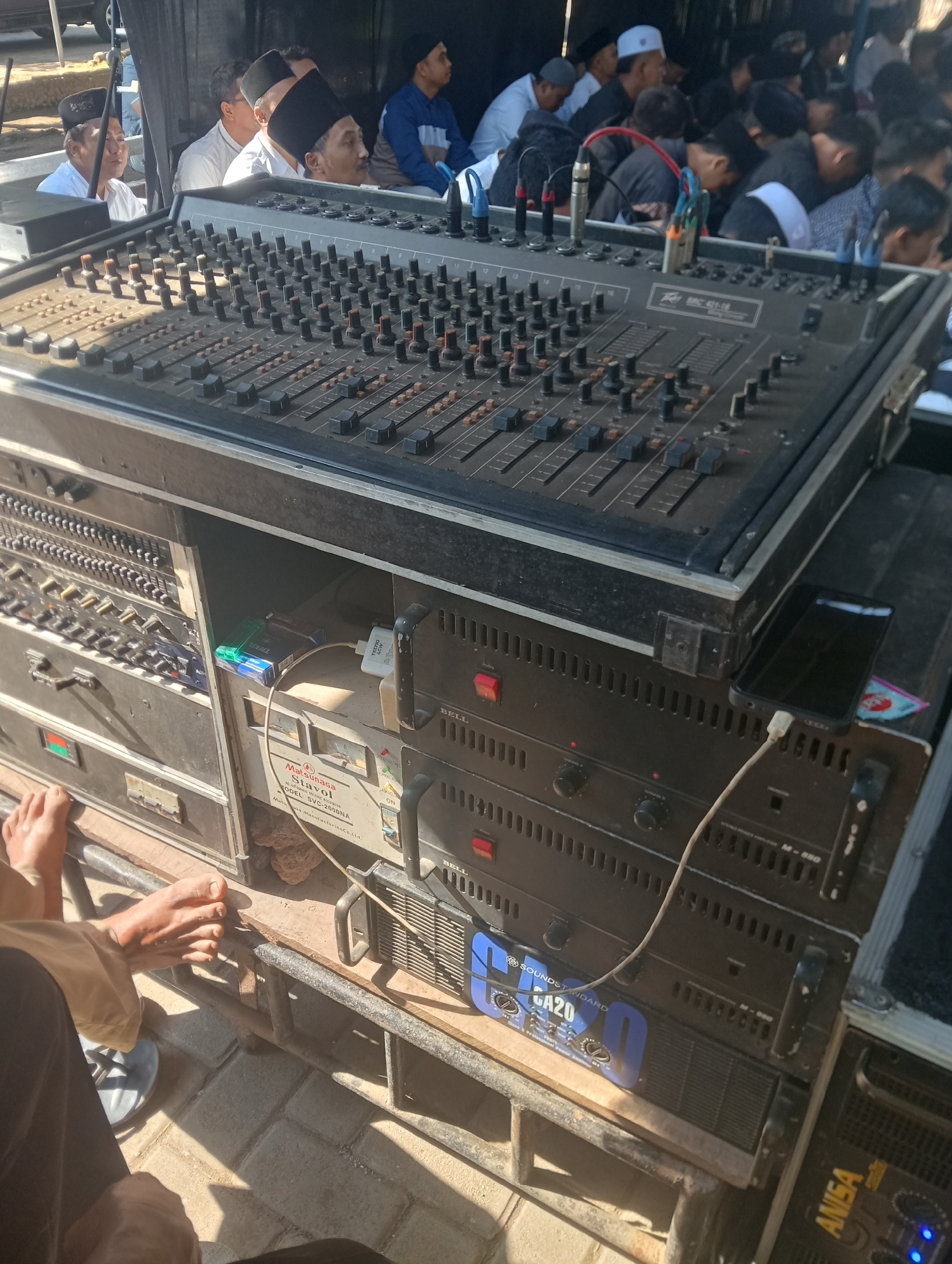 This is a medium shot of an outdoor event in Buduan, Indonesia, focused on audio equipment. In the foreground, a large audio mixing board with numerous sliders and knobs is prominently displayed. Below the mixer, several audio amplifiers are stacked, some with visible labels such as "SOUNDSTANDARD GA20" and "M-880". A black smartphone is charging on top of one of the amplifiers, connected to a power adapter. To the left, a person's bare feet are visible, along with a sandal. In the background, a group of men wearing traditional Islamic skullcaps and head coverings are gathered, some appearing to be conversing or listening. The environment appears to be a shaded area, possibly under a tent or canopy, with a tiled or paved ground. The lighting suggests it is daytime. The overall scene depicts a sound setup for a local gathering or event.