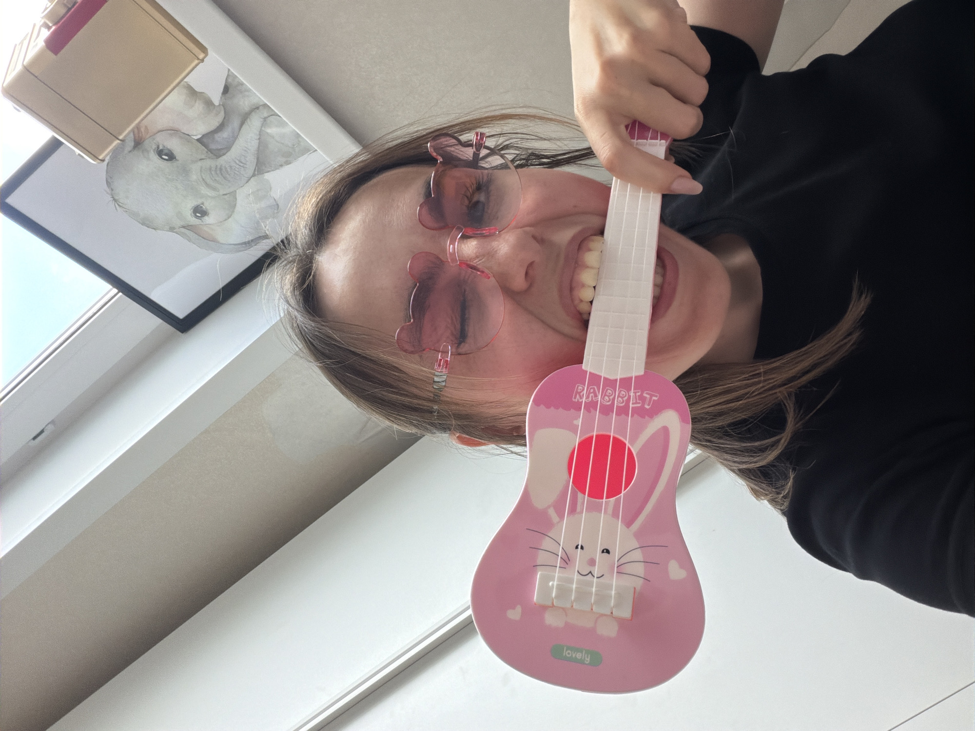 A woman with fair skin and shoulder-length brown hair is holding a pink toy ukulele with a rabbit design on it close to her face. She is wearing pink sunglasses shaped like bears. She is smiling broadly, showing her teeth. She is wearing a black shirt.
In the background, there is a window with a white frame and a framed picture of a baby elephant hanging on the wall above the window. There is a tan object (possibly a box) above the picture of the elephant. The wall is white.
The image does not contain enough detail to definitively identify the location as Izhevsk, Russia. It appears to be an indoor, personal photograph.