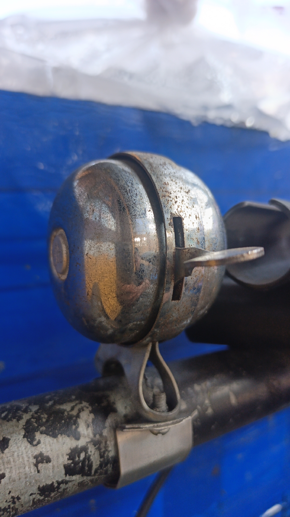 A close-up, eye-level, medium shot focuses on a rusty, chrome bicycle bell mounted on the handlebars of a bicycle. The bell is spherical and metallic, with visible signs of wear and tear, including pitting and discoloration. A small metal lever protrudes from the side of the bell. The handlebars are dark and appear worn, with some chipped paint. The background is a vibrant, out-of-focus blue, possibly a painted surface or fabric, with a blurred white element at the top suggesting a distant object or light source. The scene is static and appears to be an outdoor shot, possibly in an urban environment, though no specific landmarks in Banjarmasin, Indonesia, are identifiable from the image.
