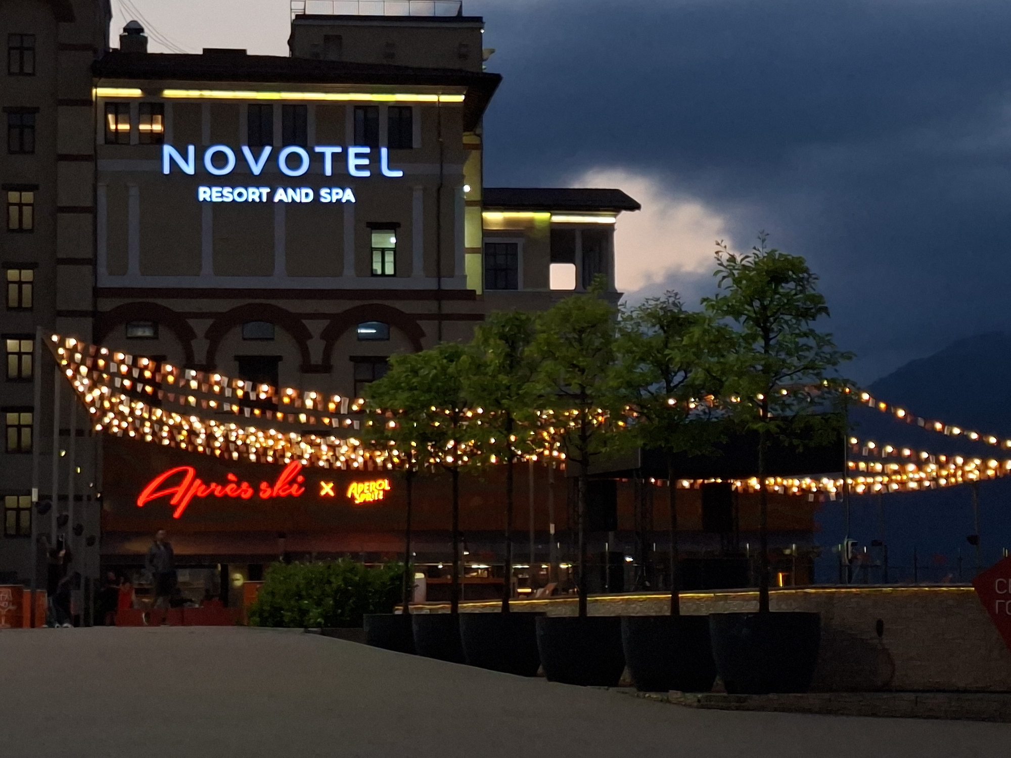 The image shows the exterior of a Novotel Resort and Spa at dusk. The hotel has a light brown facade with white trim and arched windows on the lower levels. A large illuminated sign displays "NOVOTEL" in blue letters, with "RESORT AND SPA" below it in smaller white letters. Above the hotel, a dark blue sky with some lighter clouds is visible.
In front of the hotel, strings of small, glowing lights are draped across a balcony or overhang. Below this, a red neon sign reads "Après ski" with a symbol and "APEROL SPRITZ" next to it. Several young trees in large dark planters are lined up in front of the building, their leaves illuminated by the string lights.
In the foreground, there is a paved area and some people are visible walking around. One person in a dark jacket and lighter pants is standing in the middle ground, facing away from the camera. Others are present further in the background, but are less distinct. The overall setting appears to be an outdoor area associated with the hotel, possibly a dining or entertainment space. The atmosphere is illuminated by artificial lights, contrasting with the darkening sky.