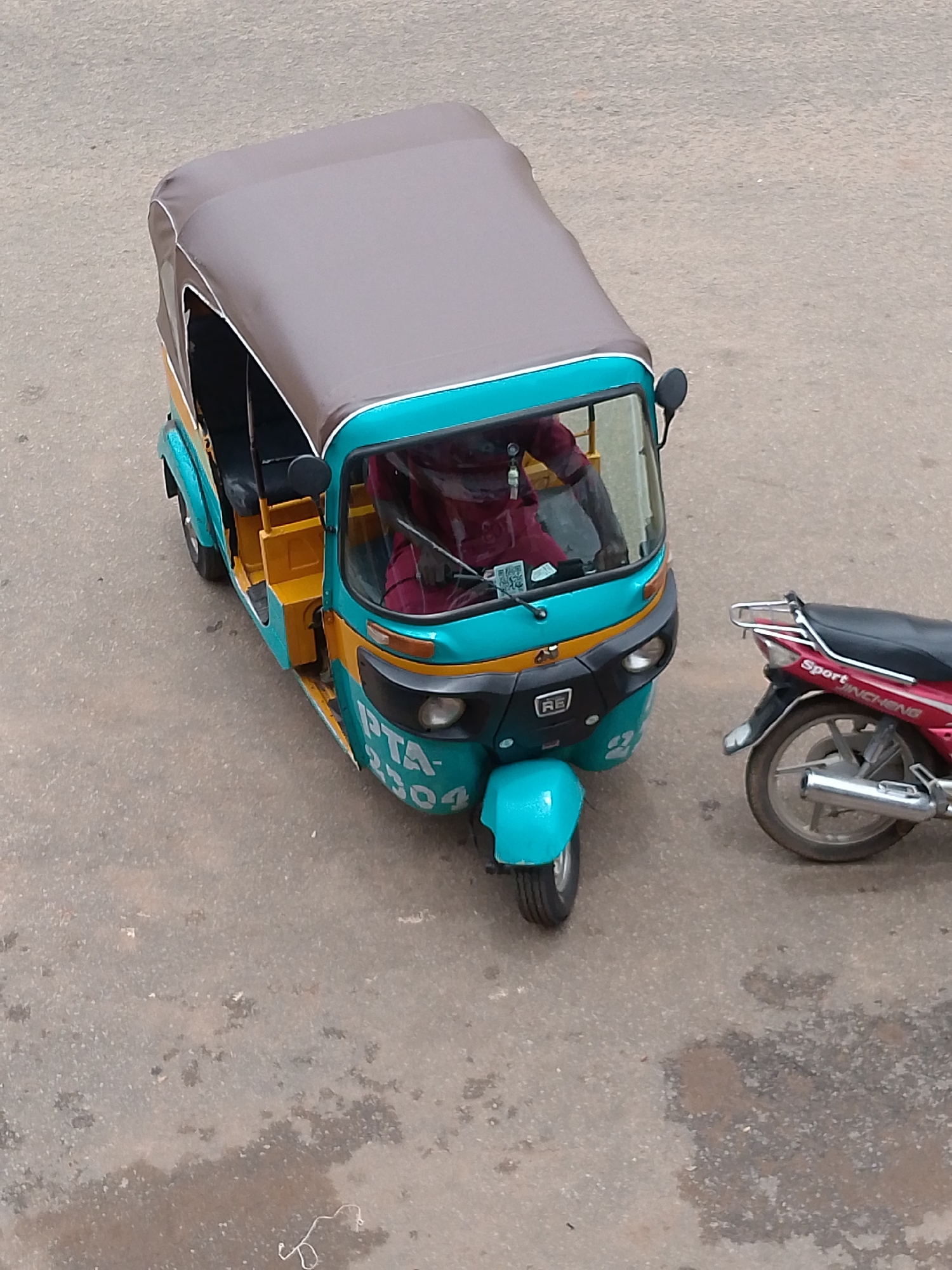 An aerial, eye-level shot shows a teal and yellow auto-rickshaw with a brown canvas roof parked on a grey asphalt road, next to a red and black motorcycle. The auto-rickshaw has a license plate that reads "PTA-2504". A person wearing a red shirt is visible inside the auto-rickshaw, sitting in the driver's seat. The motorcycle, parked to the right of the auto-rickshaw, has the word "SPORT" written on its side in white lettering. The road surface appears worn and has some dark patches that may indicate water or oil stains. The background is out of focus, suggesting a street environment.