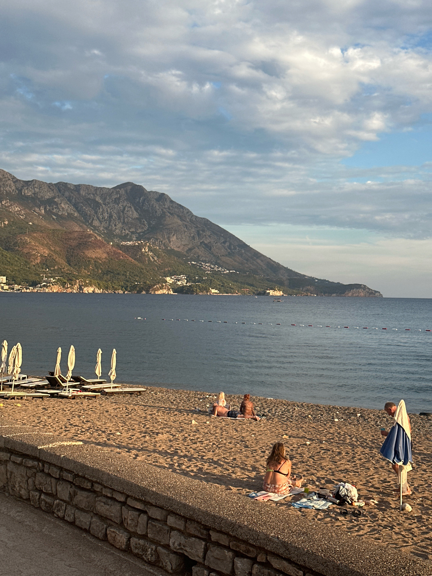This image depicts a serene beach scene in Boreti, Montenegro, under a partially cloudy sky. The foreground features a sandy beach with a stone wall bordering a walkway. Several beach chairs and closed umbrellas are neatly arranged on the sand. In the middle ground, a few people are relaxing on the beach, some sitting and others lying down. A woman in a bikini is seated on a towel, and a man stands near a partially open umbrella, holding a cup. In the background, a calm sea stretches towards a mountainous coastline dotted with buildings and vegetation. The mountains are rugged and cast shadows as the sun begins to set, with warm light illuminating their slopes. The overall atmosphere is peaceful, capturing a typical late afternoon at a coastal resort.