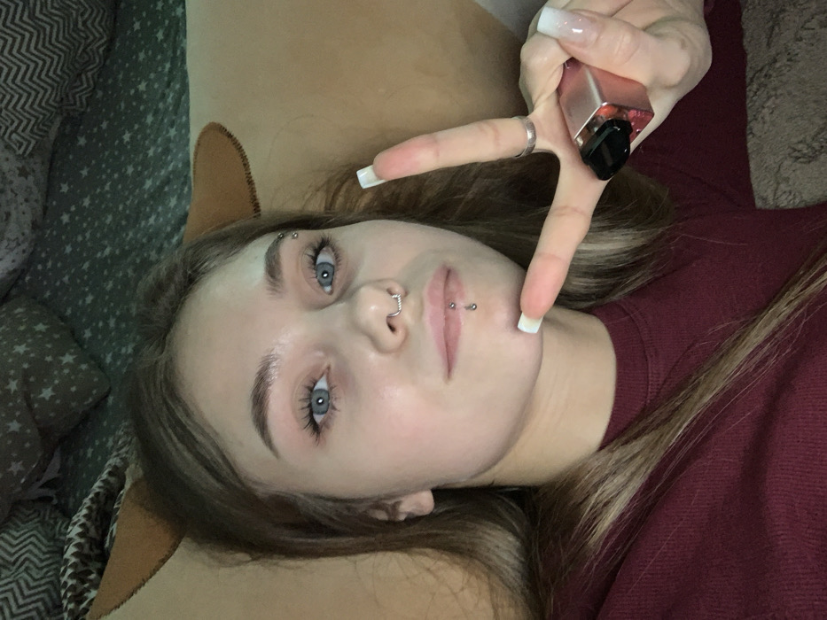 The image displays a young Caucasian female, appearing to be in her late teens or early twenties, situated in an indoor residential environment within the city of Фоки, Russia. She is depicted from the chest up, seemingly reclining. The subject has light brown hair, blue eyes, and visible facial piercings, including a silver circular septum ring, a silver barbell in her left eyebrow, and a silver vertical labret piercing on her lower lip. She is dressed in a maroon long-sleeved top. Her left hand is raised, forming a V-sign with her index and middle fingers, displaying long, square-shaped fingernails with a white ombré manicure. A silver-colored ring is visible on her left ring finger. Gripped between her left ring and pinky fingers is a small, metallic, rose-gold colored object with a black top, consistent with a personal vaping device or cosmetic product. The background consists of a light brown pillow behind her head and a dark green pillow with a light star pattern to the upper left. The subject is looking directly at the camera with a neutral facial expression.