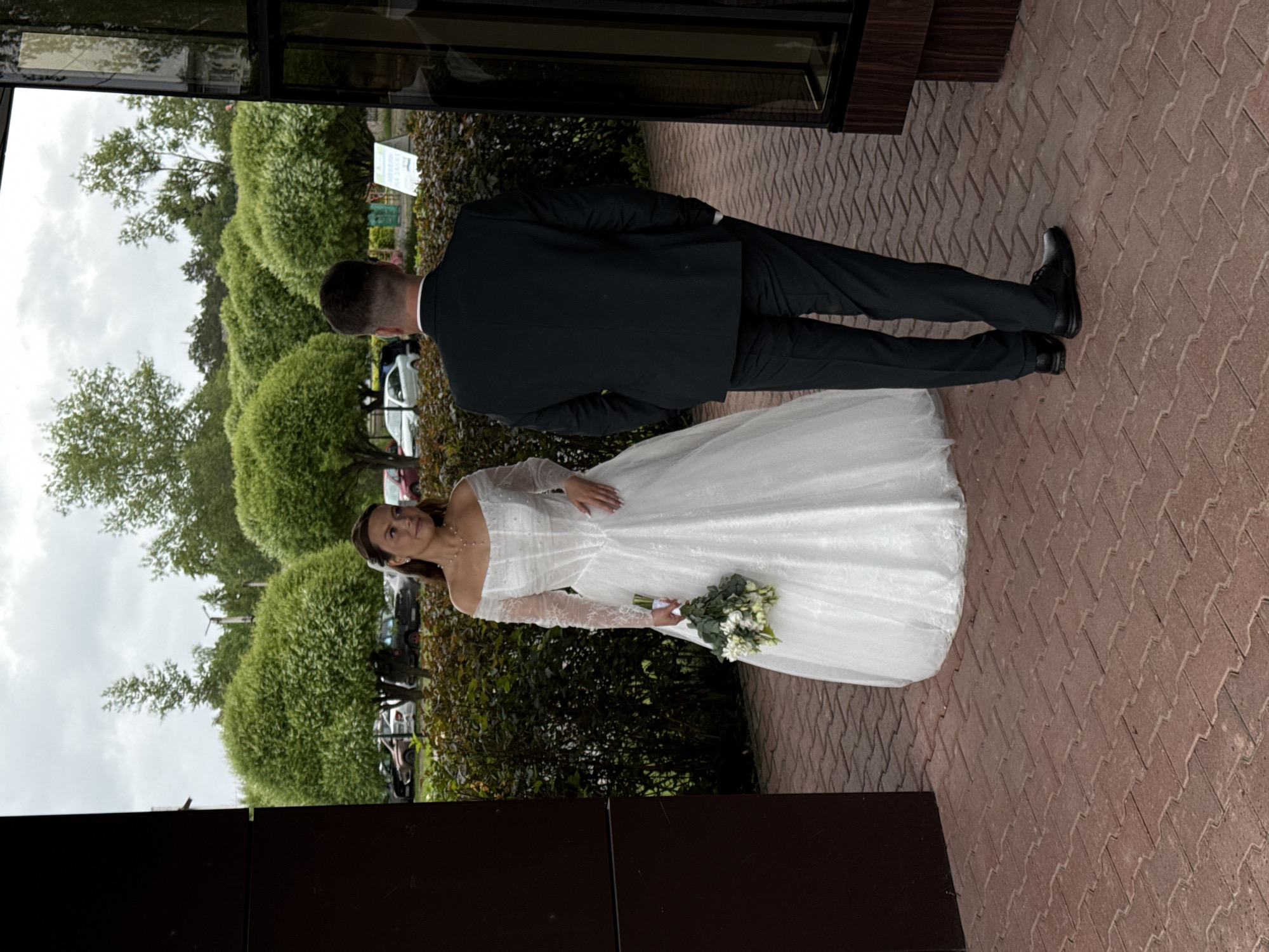The image, correctly oriented, displays a wedding couple outdoors under an overcast sky. In the foreground, a man, presumably the groom, stands with his back to the viewer, dressed in a dark suit and black dress shoes. To his left and slightly forward, a woman, the bride, faces the viewer with a subtle smile. She wears a white, off-the-shoulder wedding gown with long lace sleeves and holds a bouquet of white flowers with green foliage in her left hand.
They are positioned on a pathway paved with reddish-brown interlocking bricks arranged in a herringbone pattern. The background features lush green landscaping, including neatly trimmed, rounded bushes and taller trees. Partially visible vehicles can be discerned behind the greenery in the distance. A dark-colored building with reflective panels is present on the right side of the frame.
A green sign in the background behind the groom contains blurry, partially visible Cyrillic text. The scene captures a formal wedding event, and the provided context places the location in Chaikovsky, Russia.
