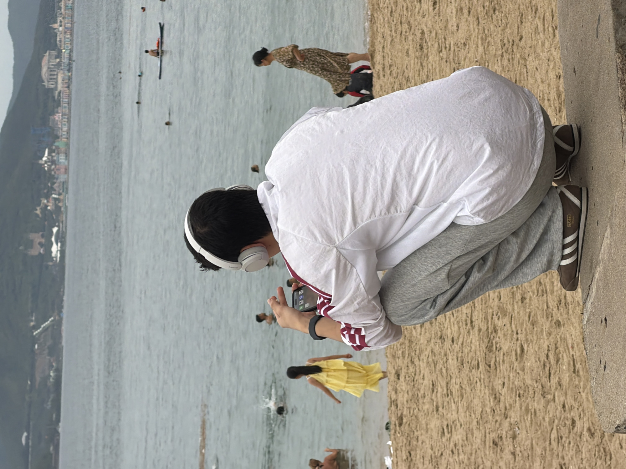 A person wearing headphones and a white jacket with red stripes is crouching on a concrete ledge overlooking a beach. The person is holding a smartphone in their right hand. In the background, people are swimming and wading in the ocean. A young girl in a yellow dress is standing near the water's edge. A woman in a patterned dress is also near the water. The beach sand is visible in the foreground and middle ground. In the far background, a city skyline with buildings and hills can be seen. The location is identified as Nha Trang, Vietnam.