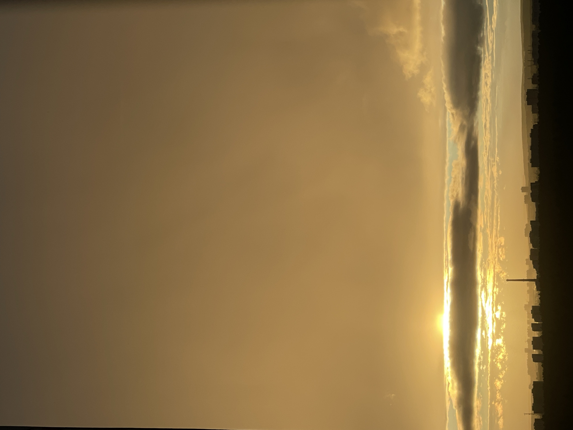 This is a vertical, wide-angle photograph of a sunset over a cityscape in Perm, Russia. The sun is setting on the right side of the frame, casting a warm, golden light across the sky. The sky is filled with a variety of clouds, some of which are illuminated by the setting sun, while others are darker and more dramatic. A distinct cloud formation, appearing as a long, dark band, stretches vertically through the center of the image, with the sun peeking through its edges. In the distance, on the right side of the frame, the silhouettes of buildings and a tall industrial chimney are visible, indicating an urban environment. The overall mood of the photograph is serene and awe-inspiring, with the dramatic lighting and cloud formations creating a sense of grandeur. The time of day is clearly sunset, given the sun's position and the warm hues of the sky.