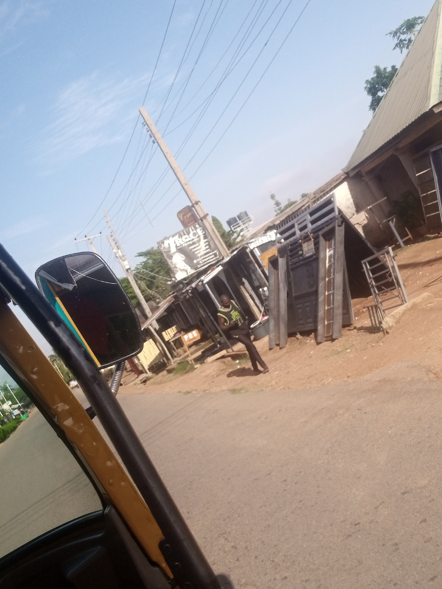 The image captures a street scene in Gura Top, Nigeria. In the foreground, a portion of a yellow and black vehicle, likely an auto-rickshaw or tuk-tuk, is visible, including its side mirror. The mirror appears cracked. A person wearing a green and black sleeveless shirt and dark pants is standing on the dirt roadside, looking down and to their right. Behind the person, several metal structures, possibly gates or doors, are leaning against a building. The building has a corrugated metal roof. Power lines are strung across the sky, supported by wooden poles. A sign with text, including the word "TRAGE", is attached to one of the poles. The ground is a mixture of dirt and asphalt. The overall environment suggests a commercial or workshop area.