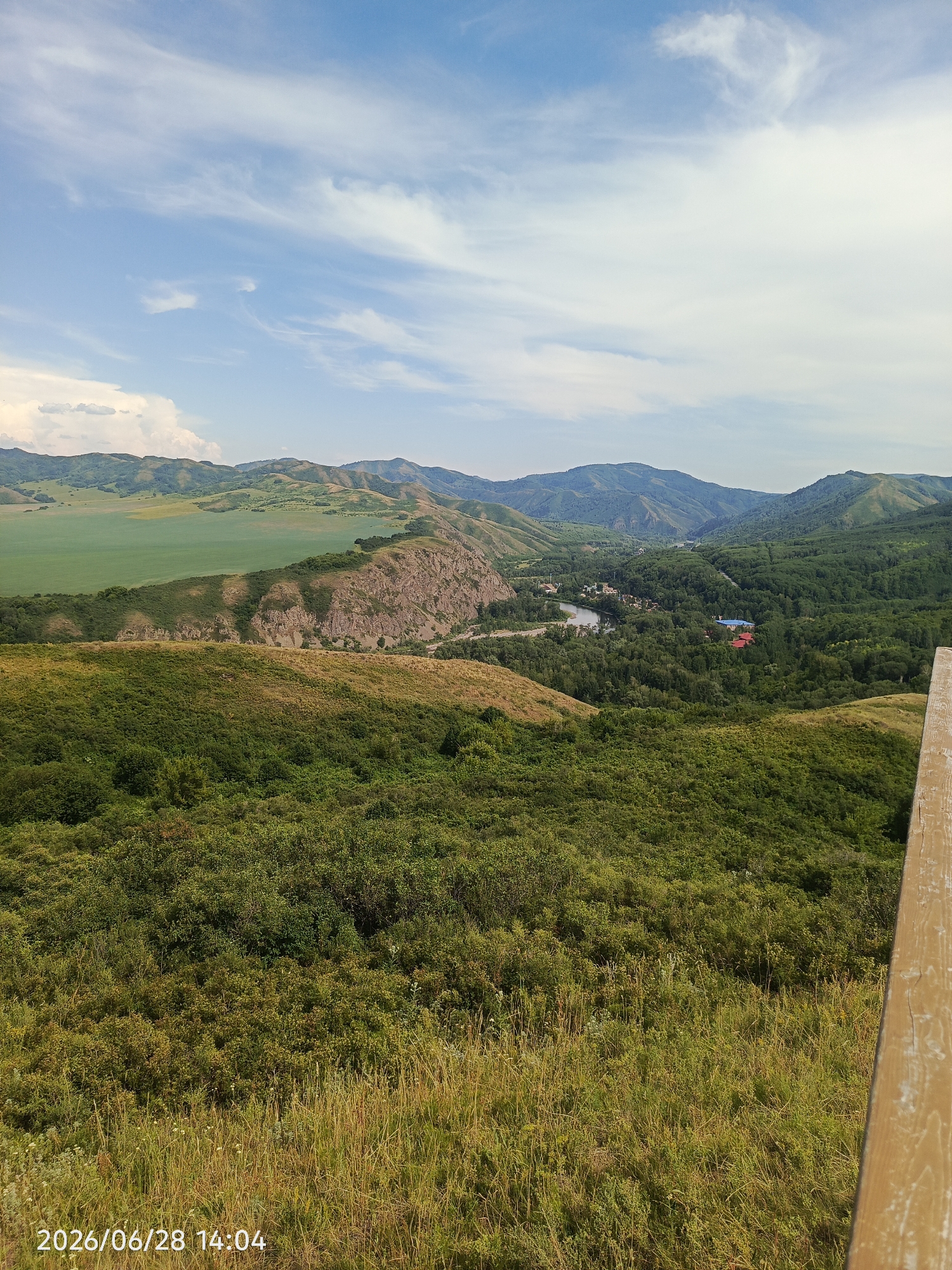 The image shows a vast, green landscape with rolling hills and a valley. In the foreground, there is a grassy hillside with tall dry grass and lush green bushes. Further in the distance, a rocky outcrop juts out from a greener hillside. Beyond this, a valley opens up, filled with dense green trees. A river winds through the valley, with a few buildings visible on its banks, including a blue-roofed building and a red-roofed building. In the far distance, a series of hazy, green mountains stretches across the horizon under a blue sky with scattered white clouds.
The scene suggests a natural, rural setting. The time of day appears to be afternoon, indicated by the brightness of the sky and the shadows cast. The weather seems pleasant, with a clear blue sky and only a few clouds. The location is identified as "Тарханка ауылдық округі, Kazakhstan" in the prompt.
No people are visible in the image. The focus is entirely on the natural scenery and the remote settlement in the valley. The overall impression is one of tranquility and natural beauty.
The visible text in the bottom left corner reads "2026/06/28 14:04", indicating the date and time the photograph was taken. A wooden railing is partially visible on the right side of the frame.