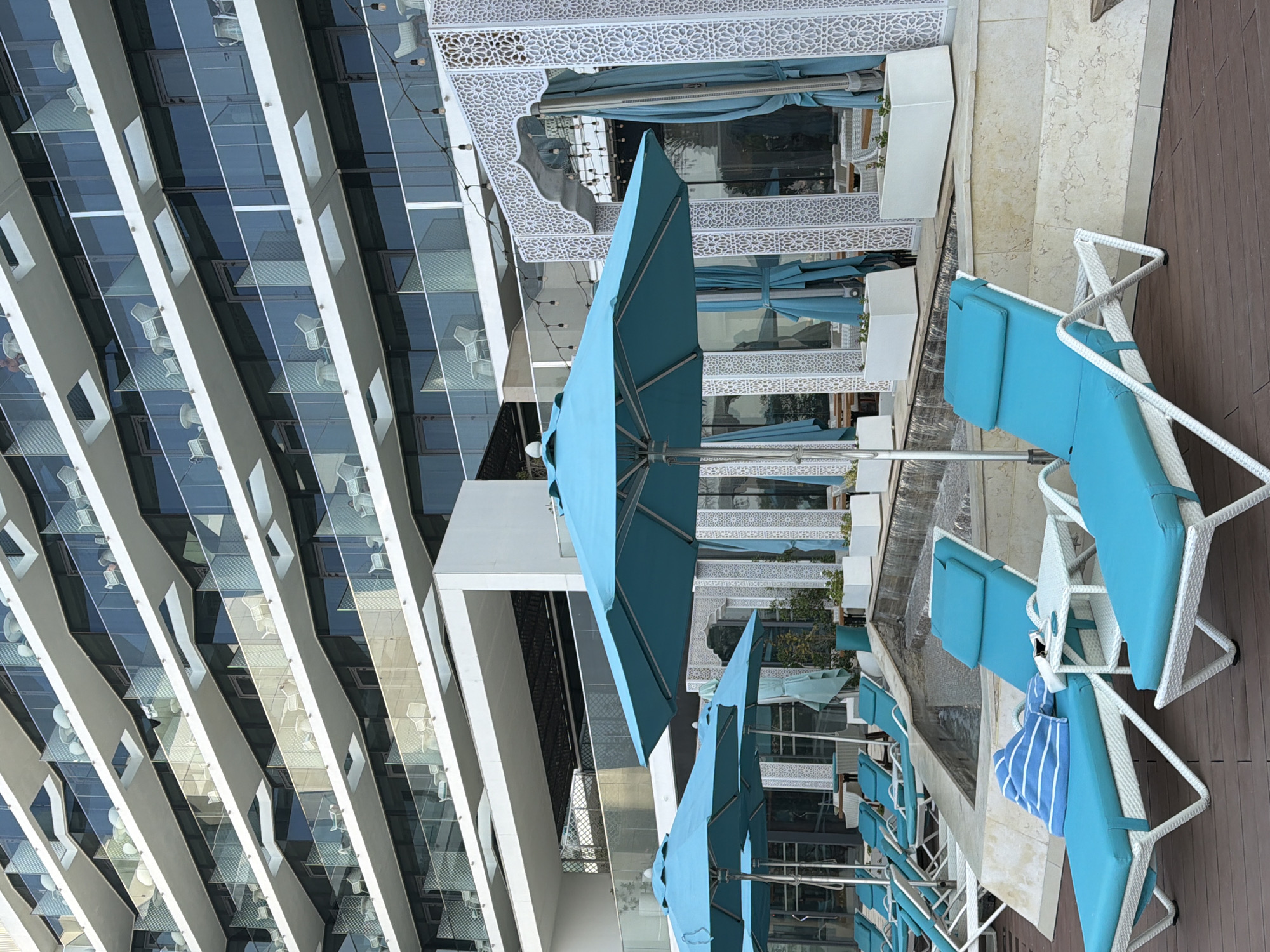 The image shows a resort scene in Dubai, United Arab Emirates. In the foreground, there are several bright blue lounge chairs with white frames, arranged on a wooden deck. Some of the chairs have blue and white striped towels draped over them, and one has a small white side table next to it. Behind the lounge chairs, there are several large, teal umbrellas providing shade. Beyond the umbrellas, a tiered water feature with small waterfalls is visible. In the background, a modern, multi-story building with white balconies and glass railings dominates the scene. The building appears to be a hotel or resort. Intricate white latticework structures with decorative arches are also present, suggesting an outdoor dining or lounge area. The overall impression is one of a luxurious, relaxing environment.