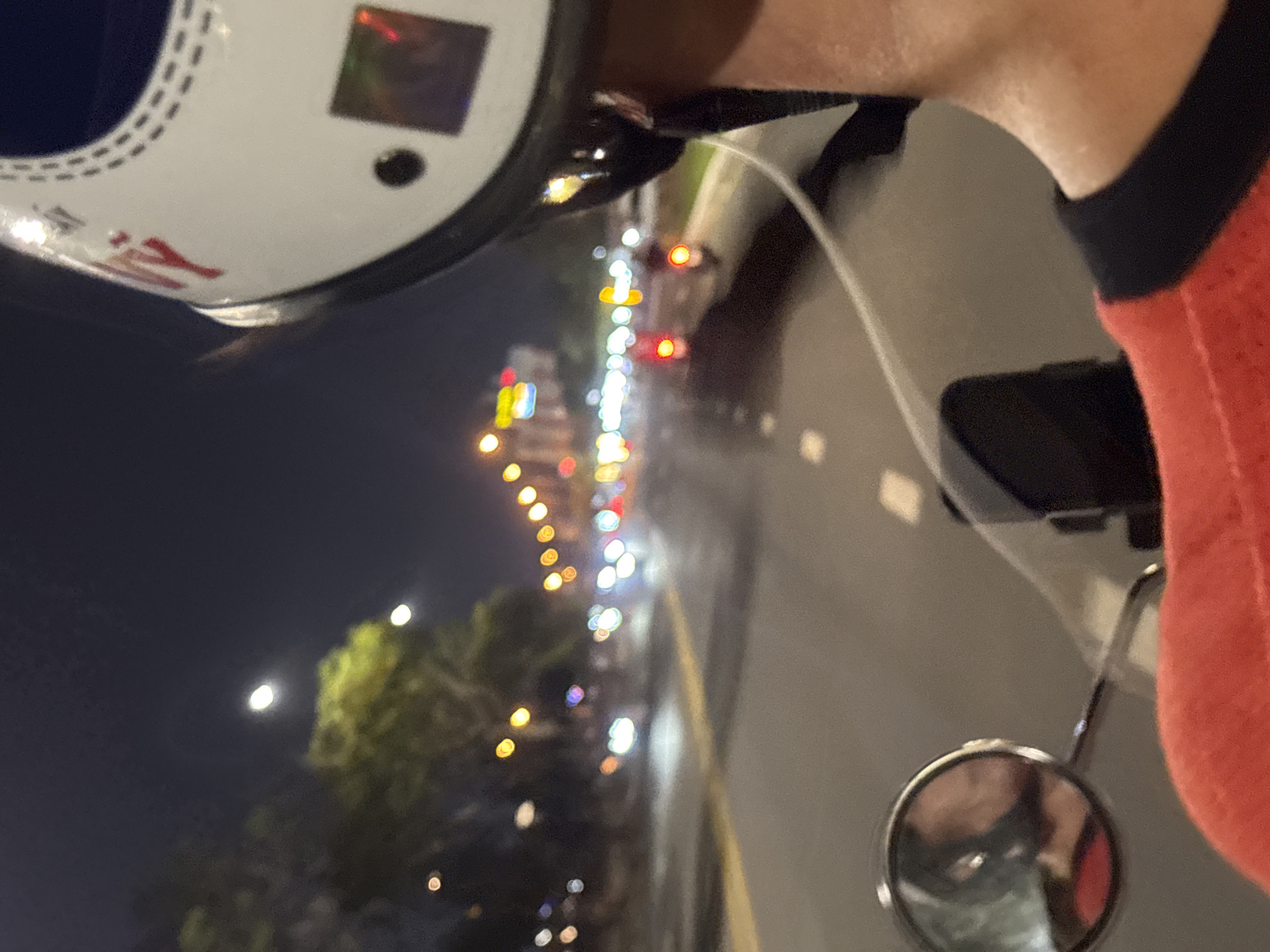 A first-person perspective image, taken at night, shows a view from a motorcycle, likely on a street in Đà Nẵng, Vietnam. The rider is wearing a helmet, part of which is visible in the upper left corner. A black neck brace is seen near the rider's right shoulder. The motorcycle's side mirror is in the lower right corner, reflecting a blurred image of the rider's face, presumably wearing a mask.
The road stretches ahead, lined with blurred city lights suggesting traffic. Some trees can be seen to the left of the road. A bright full moon is visible in the sky. The image shows a low angle and blurred effect which indicates the vehicle is in motion.