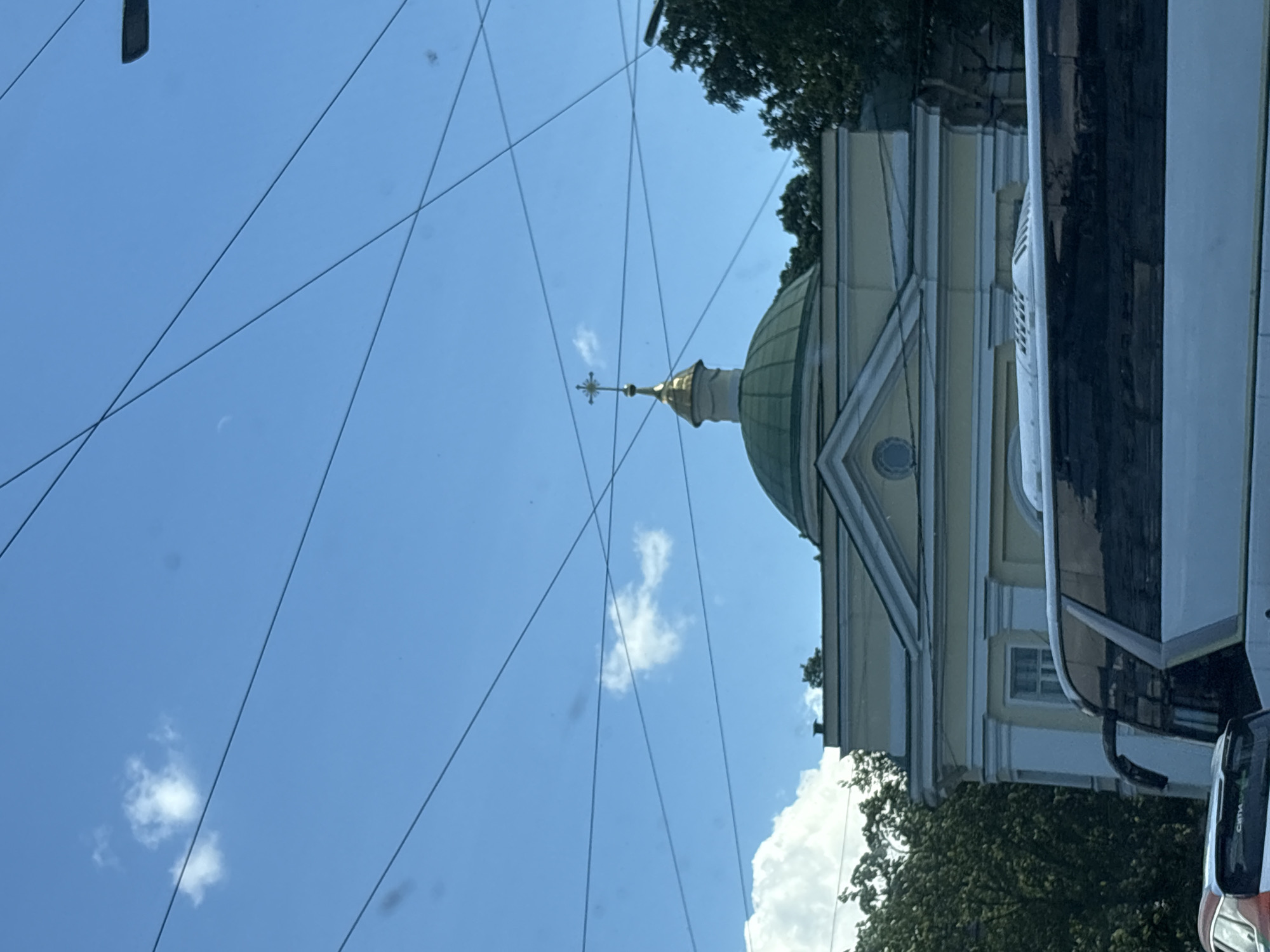 The image shows a building with a green dome and a golden cross on top, likely a church or cathedral, in Saint Petersburg, Russia. The building has a light yellow facade with white architectural details. A large white bus is partially obscuring the lower part of the building. The sky is blue with some white clouds and overhead power lines crisscross the frame. Trees are visible on the right and left sides of the building, suggesting an urban environment. The image appears to be taken from a street level, possibly from another vehicle given the presence of the bus.