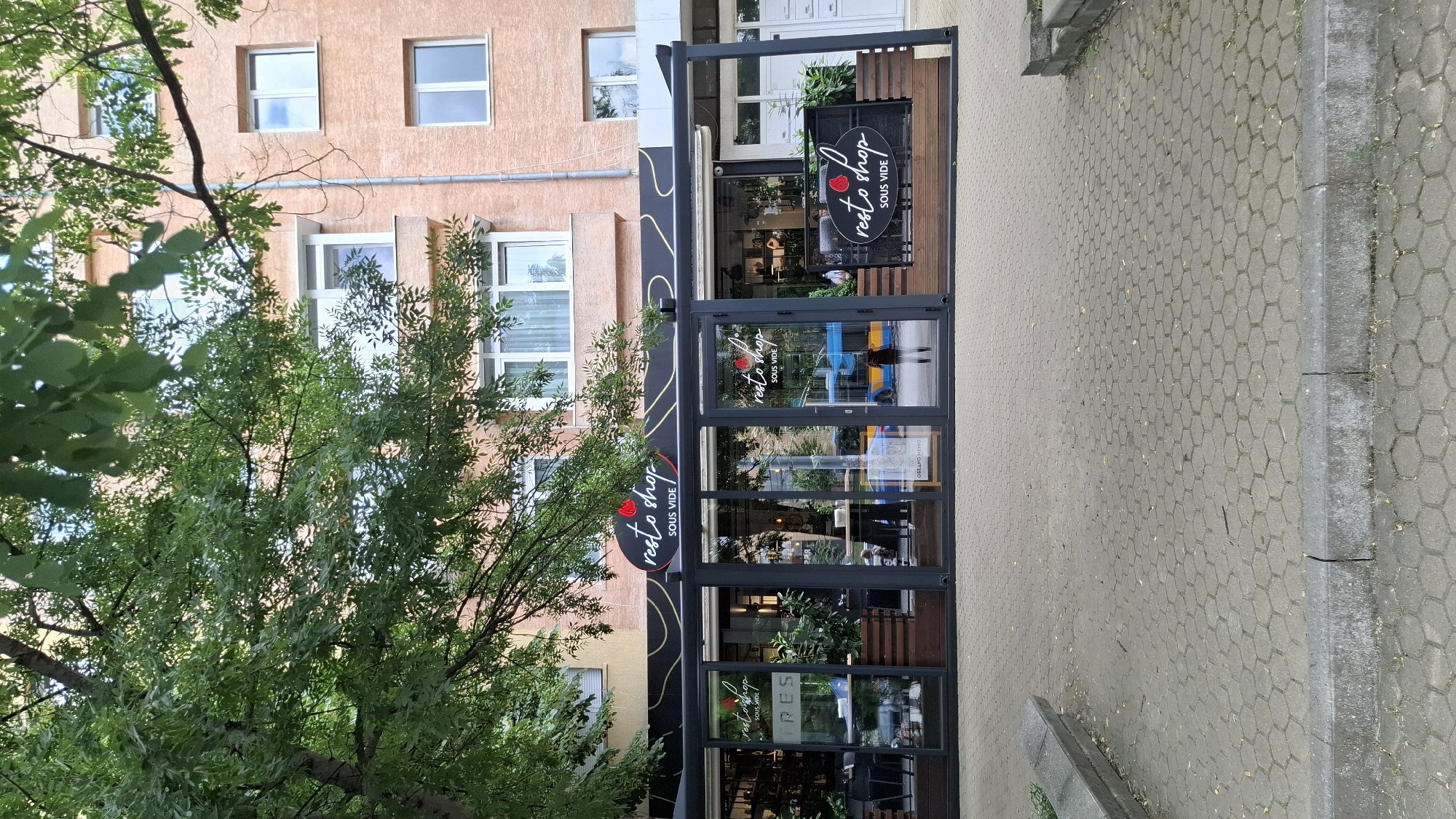 A street-level, eye-level shot shows the exterior of a modern restaurant and shop with a covered outdoor seating area. The building facade is painted a light orange color and features multiple windows. A large tree with green leaves is in the foreground, partially obscuring the view of the building and the restaurant's signage, which reads "resto shop SOUS VIDE" in a stylized font. The restaurant has large glass doors and windows, revealing tables and chairs inside. Wooden planters with greenery are placed along the front of the covered seating area. The sidewalk in front of the establishment is paved with hexagonal gray tiles. In the background, a blue bus is visible through the glass of the restaurant. The image was taken in Sofia, Bulgaria.