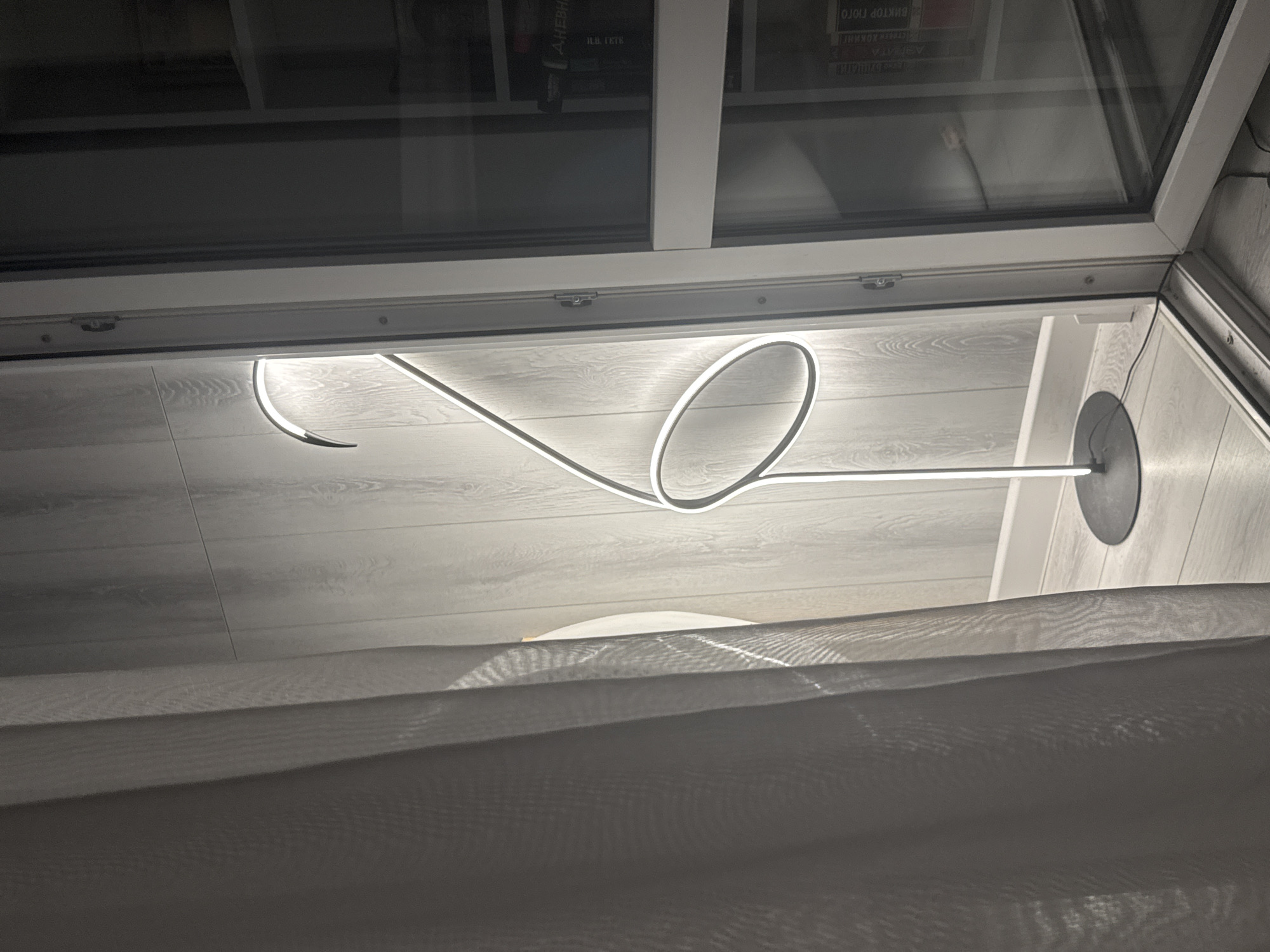 The image appears to be taken from a low angle, looking up towards a ceiling with a skylight. The ceiling is paneled with light-colored, horizontally oriented planks. A modern, minimalist lamp is mounted on the ceiling; it consists of a thin, illuminated tube that bends into a circular shape before extending to a circular base affixed to the ceiling. A white curtain hangs down in the foreground, partially obscuring the view. The skylight, framed in white, reveals glimpses of bookshelves with books in the background. The location cannot be determined from the image.