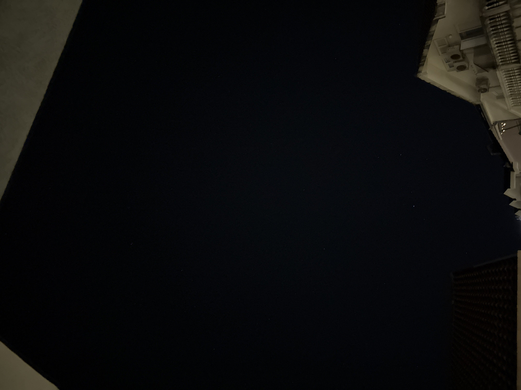 The image captures a night scene from Budva, Montenegro, likely from an elevated vantage point such as a balcony. The sky is a deep, dark blue, speckled with numerous small stars, indicating a clear night. In the foreground, the dark red tiled roof of a building is visible in the lower left corner. To the right, a white building with architectural details such as windows, air conditioning units, and a balcony with decorative railings is partially visible. The overall impression is one of a quiet, residential night in a coastal city.