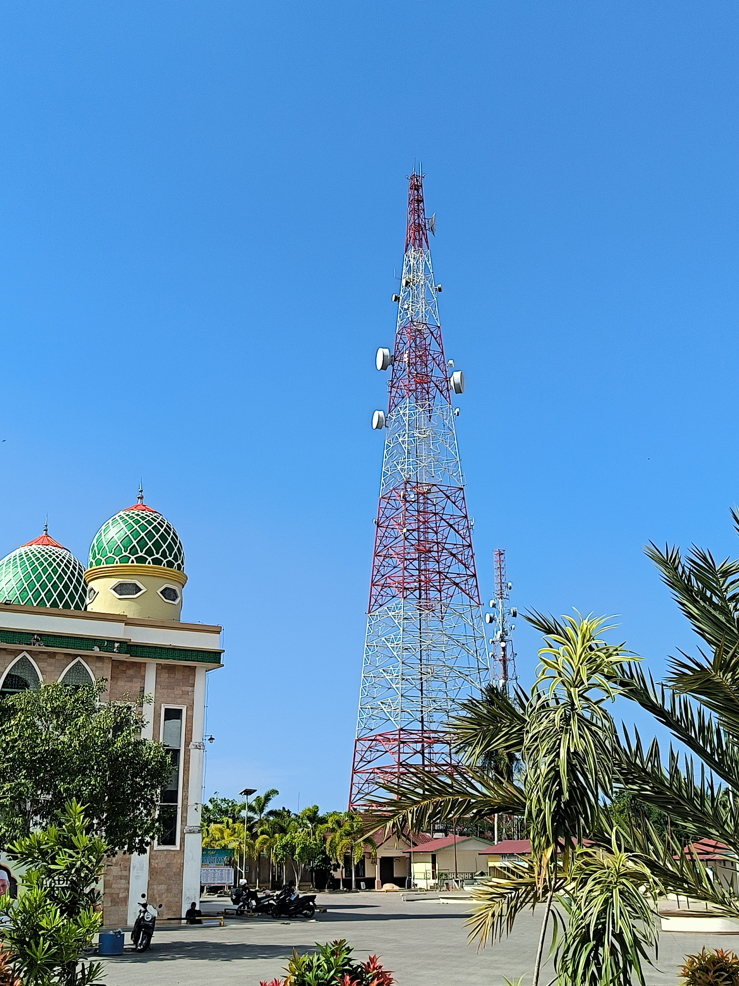The image shows a landscape in Titibaro, Indonesia. A tall, red-and-white telecommunications tower dominates the background, standing against a clear blue sky. To the left, a mosque with two green, dome-shaped roofs is partially visible. In the foreground, various trees and shrubs, including palm trees, add to the natural setting. Below the tower, there is a building behind the palm tree. In front of the mosque, there are several motorcycles parked. A person is sitting on a bench. Another telecommunications tower is partially visible near the main tower. The ground appears to be a paved area.