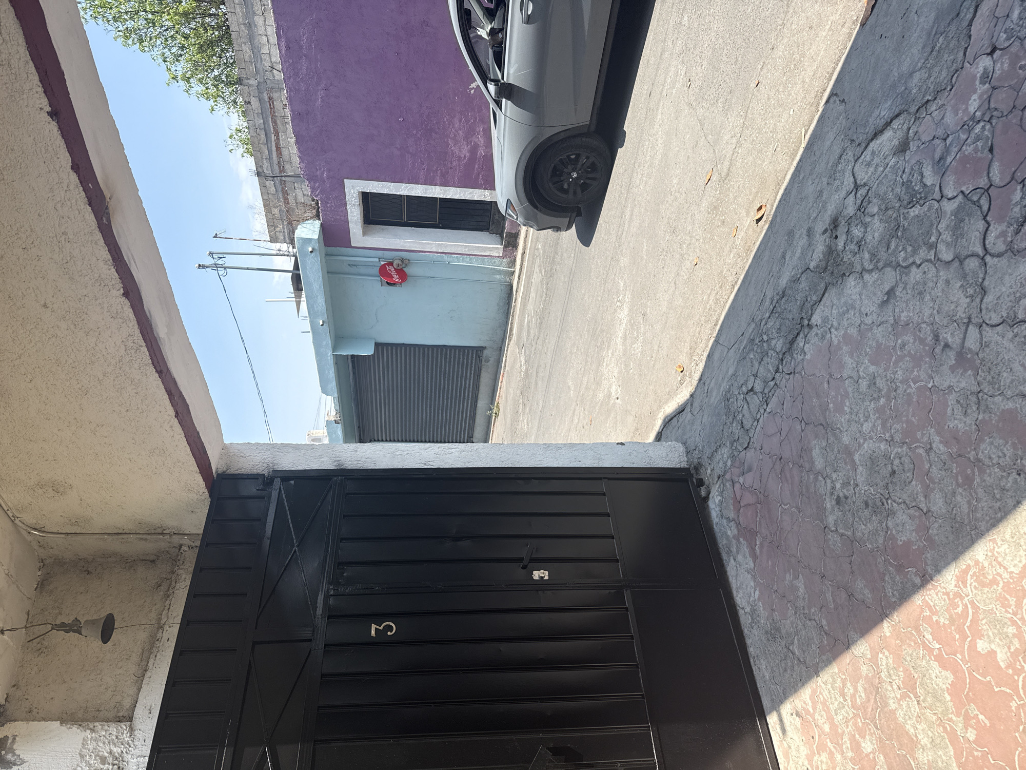 The image is a vertical shot depicting a sunlit outdoor scene, likely a street or alleyway in Xochitepec, Mexico. The composition suggests the camera was held horizontally and the image rotated 90 degrees clockwise.
In the lower portion of the frame, a large black metal gate is prominent, featuring horizontal panels and a latch mechanism. A silver number "3" is visible on the gate's lower right section. To the left of the gate, an overhead concrete structure, possibly a ceiling or overhang, extends into the scene. A white conical speaker or light fixture hangs from this structure, connected by wires.
The midground and upper sections of the image reveal a street bordered by buildings under a clear blue sky. A light gray SUV or crossover car is parked on a concrete road surface, oriented towards the upper right of the frame. Its front wheel and part of its side are visible, casting a strong shadow onto the ground. The road surface varies, with smooth gray concrete meeting a more textured, cracked reddish-brown and gray patterned surface. Scattered dry leaves are present on the concrete.
On the far right, a building with a vibrant purple wall stands out, featuring a white-framed window with dark security bars. Below this, a light blue-green building is visible, with a closed, metal roll-down shutter and a round, red Coca-Cola sign mounted above its entrance. Utility poles and power lines crisscross against the bright blue sky in the upper left. Green foliage from a tree can be seen peeking over the purple wall. The strong shadows and bright light indicate a clear, sunny day, likely mid-morning or afternoon.