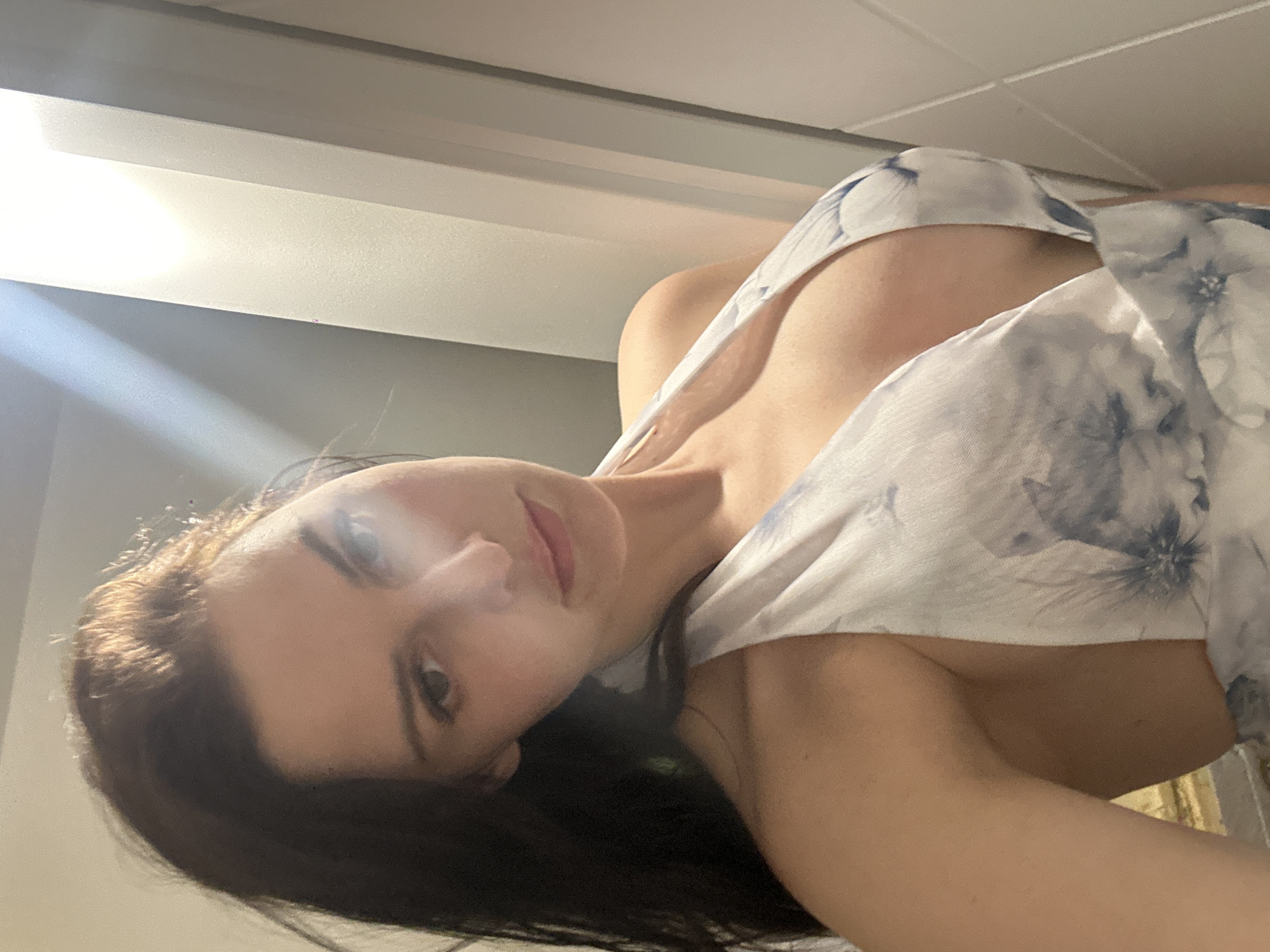 A dark-haired woman with fair skin poses for a selfie in a dress with a floral pattern. She is looking at the camera with a neutral expression. The background is out of focus and appears to be a room with a bright light source.