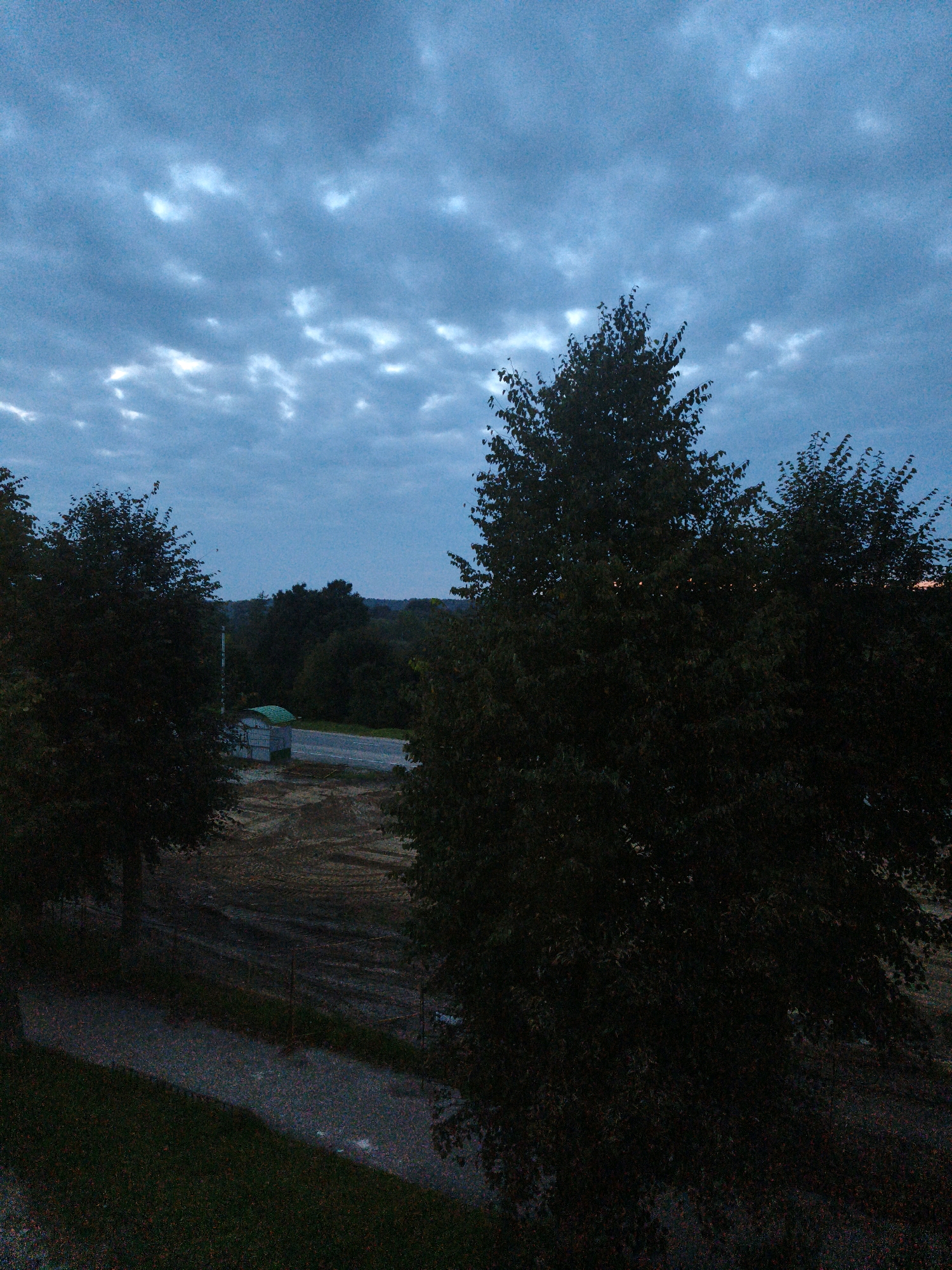 An exterior, elevated, medium shot captures a twilight scene in the village of Horki, Belarus. The sky is a deep blue, filled with scattered, elongated clouds. In the foreground, large, dark green trees dominate the left and right sides of the frame, obscuring much of the background. Between the trees, a road is visible, running horizontally across the mid-ground. To the left of the road, a small, light-colored structure with a green, sloped roof sits next to a vertical pole. The ground in front of the structure and extending towards the road appears to be tilled earth, with visible tire tracks from machinery. A narrow, gravel path or walkway is present in the lower left quadrant of the image, bordered by grass. The overall lighting suggests either early morning or late evening, with low light conditions.