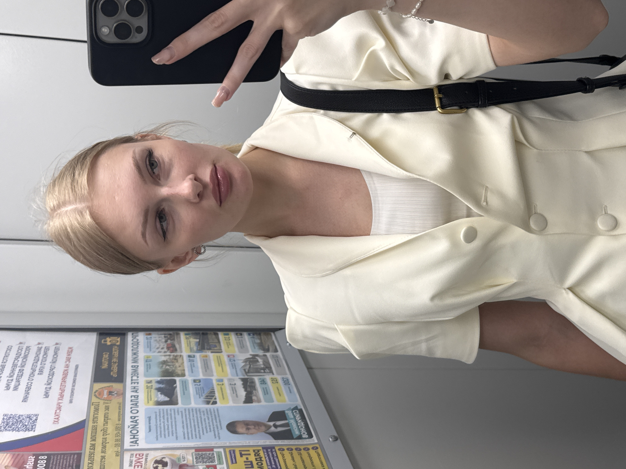 The image shows a young woman with blonde hair, wearing a cream-colored blazer over a white ribbed top, taking a selfie in an elevator. She is looking to her right, with her right arm holding a black smartphone. A black strap from a bag is visible across her body, and she is wearing a pearl bracelet on her right wrist. Behind her, on the elevator wall, are various posters, some with Cyrillic text, indicating the location is Perm, Russia. One poster shows a portrait of a man in a suit. The elevator walls are a light gray color.