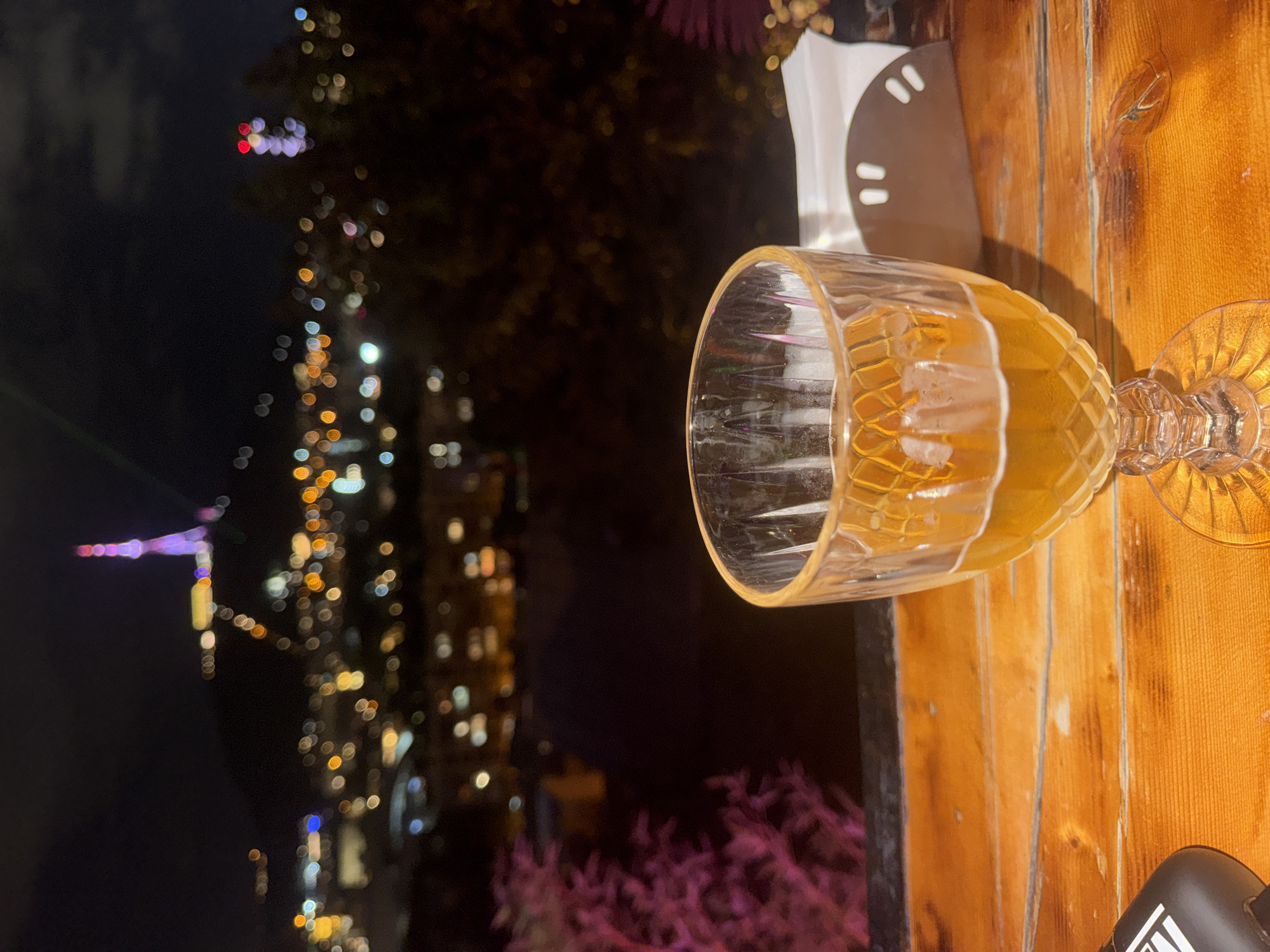 The image shows a close-up of a glass of amber-colored liquid, likely beer or wine, on a wooden table at night. In the background, the out-of-focus lights of Tbilisi, Georgia, are visible, including the Narikala Fortress and the Mtatsminda Park, featuring the Tbilisi TV tower. The scene suggests a person is enjoying a drink with a view of the city. The overall atmosphere is relaxed and evening-oriented.