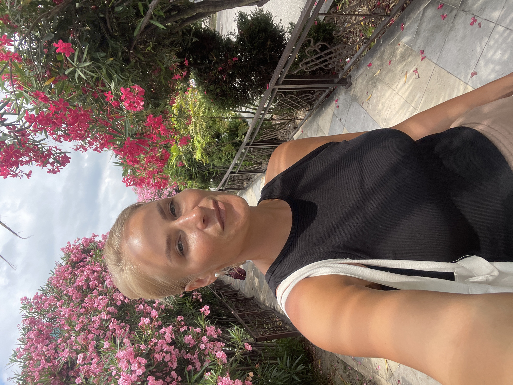 A young, blonde woman is taking a selfie outdoors in front of abundant pink oleander bushes. She is wearing a black tank top, a pearl earring, and carrying a white tote bag. Her hair is pulled back, and she is smiling at the camera. The background shows a pathway with a decorative metal railing, lined with more blooming oleander trees and various green shrubs. The ground is paved with large, light-colored tiles, and fallen pink petals are scattered across it. The sky is partly cloudy. The location is identified as Sochi, Russia.