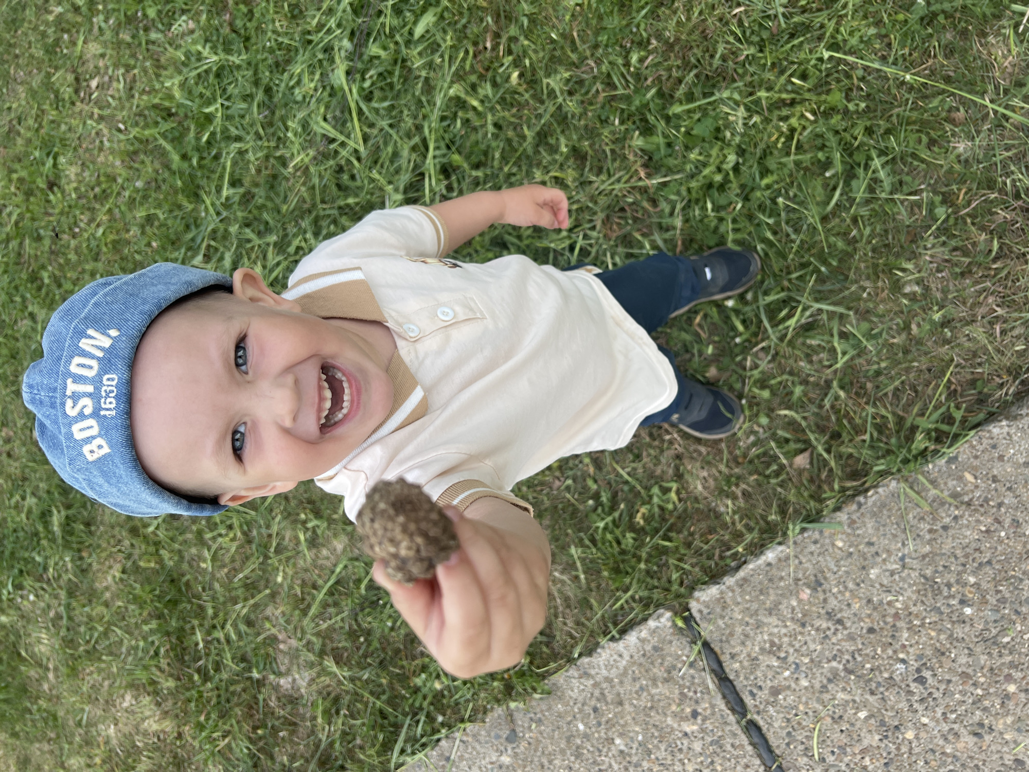 A young child stands on green grass next to a concrete sidewalk. The child is wearing a blue denim cap with the word "BOSTON" and the number "1630" embroidered on it, a tan polo shirt with tan trim, navy blue pants, and navy blue shoes. The child is holding a brown, round object in their right hand, presumably a pinecone or similar natural item. The child is smiling and looking up towards the camera. The scene appears to be outdoors during daylight hours. The location is Марковский, Russia.