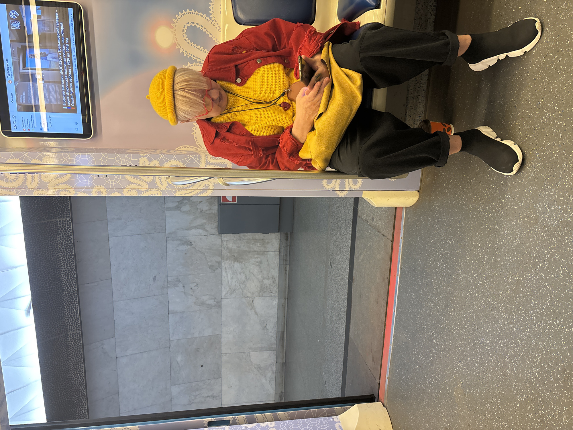 The image shows a person sitting on a seat inside a Moscow Metro train, Russia. The person is wearing a yellow beanie, red glasses, a yellow sweater, a red jacket, and black pants with black sock-style sneakers. They are holding a brown object wrapped in a yellow bag in their lap. An information screen on the left wall of the train displays "Тургеневская" station and weather information (16°C). The screen also notes an exhibition at the "РОСИЗО" and Tretyakov Gallery. The interior of the train has a modern design with grey seating, patterned wall coverings, and a grey speckled floor. Through the train windows, a platform can be seen, suggesting the train is at a station.