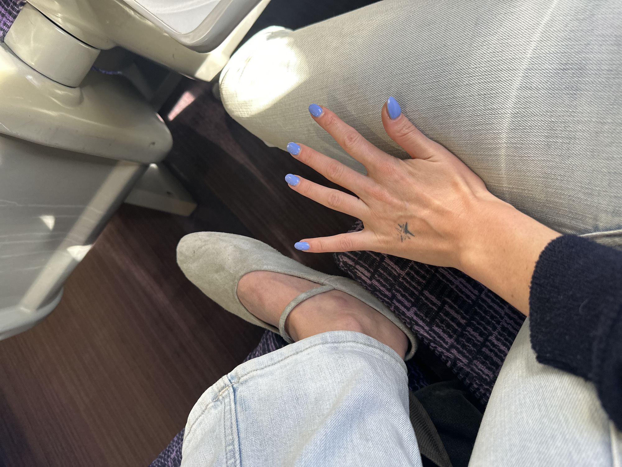 The image shows a person seated, likely on a train. The person is wearing light-wash denim jeans and a grey suede ballet flat. Their hand rests on their lap, revealing light blue nail polish and a small bird tattoo on their wrist. Part of a train seat is visible to the left. No specific location within Sevenoaks is evident from the image.