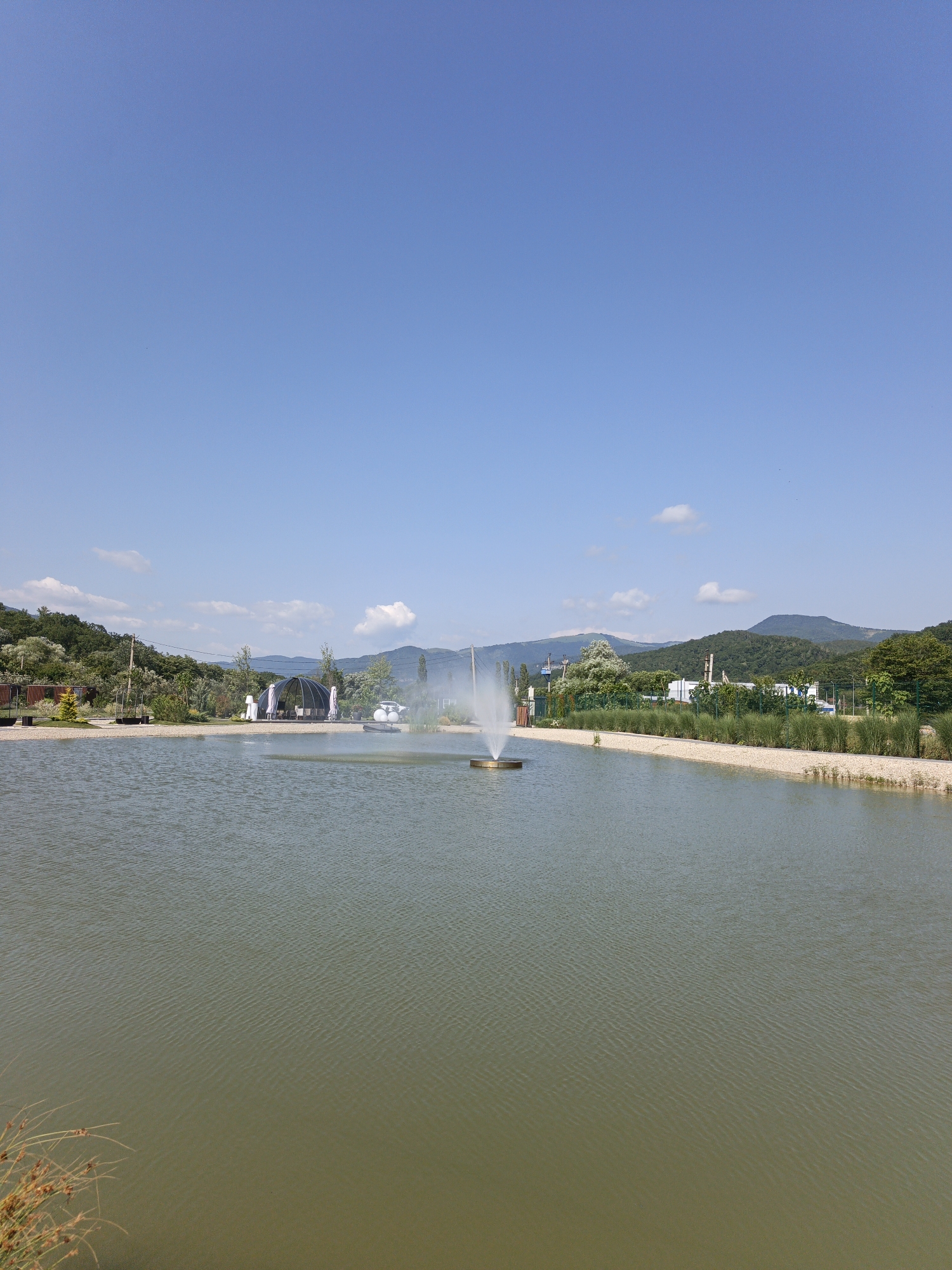 A serene, wide-angle shot captures a tranquil lake on a bright, clear day, with a prominent fountain spraying water in the middle. The lake's surface is a muted greenish-brown, reflecting the soft ripples of the water. Along the edge of the lake, a gravel pathway borders a strip of manicured green grass and low-lying shrubs. In the mid-ground, several structures are visible. To the left, a large, transparent, geodesic dome-like tent, appears to be an outdoor dining or event space, with tables and chairs set up inside. Beside it, a white car and a white tent are parked. Further to the right, industrial-looking buildings and tall structures can be seen in the distance, juxtaposed against the lush green of rolling hills and distant mountains that dominate the background. The sky above is a brilliant, cloudless blue, indicating a pleasant, sunny day. The image appears to be taken in the vicinity of the city of Pshada, Russia, a region known for its natural beauty and proximity to mountainous landscapes.
