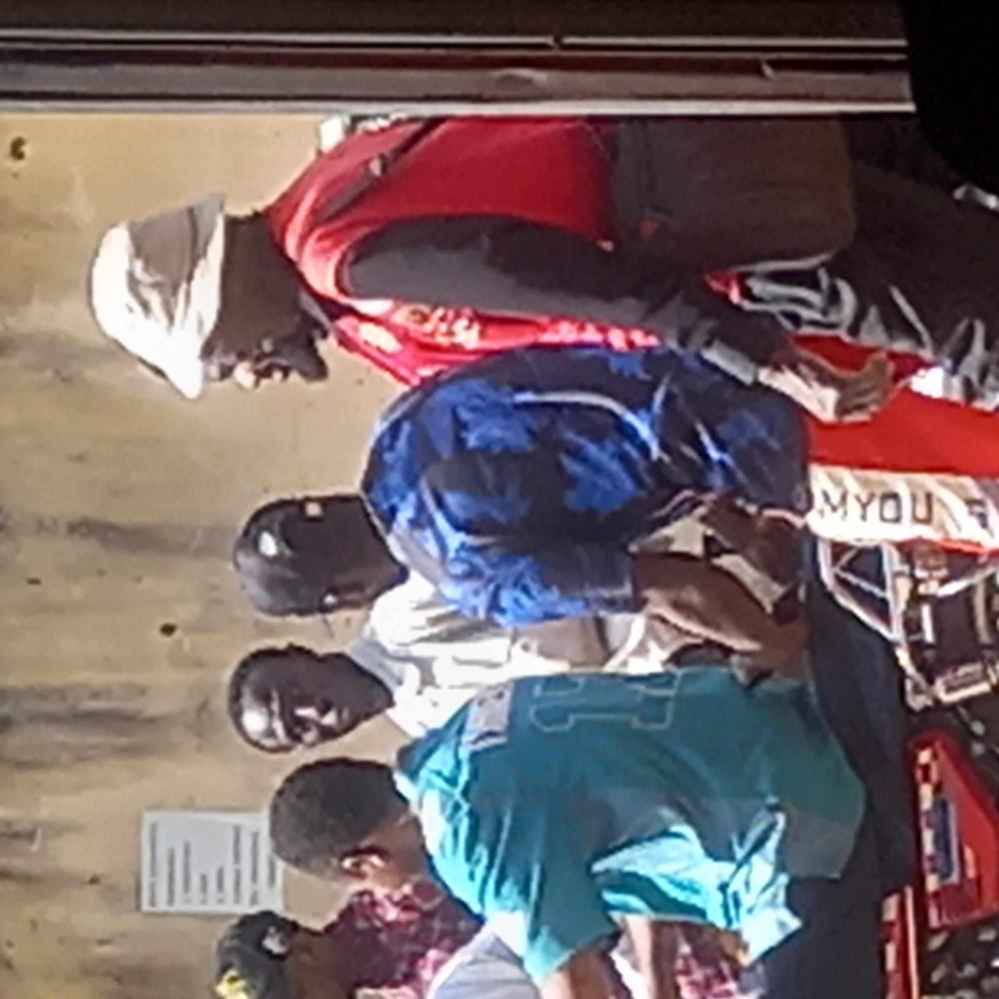 The image shows five people standing in what appears to be an outdoor setting, possibly in Dadi Riba, Nigeria, based on the prompt. The first person is wearing a white head covering, a red vest, and a black long-sleeved shirt. The second person is wearing a blue shirt with a floral pattern and a black baseball cap. The third person has on a white shirt and a black baseball cap. The fourth person is wearing a light blue shirt. The fifth person is wearing a red and purple patterned shirt and is also wearing a black baseball cap. They appear to be observing something on the ground. A sign is visible in the background.
