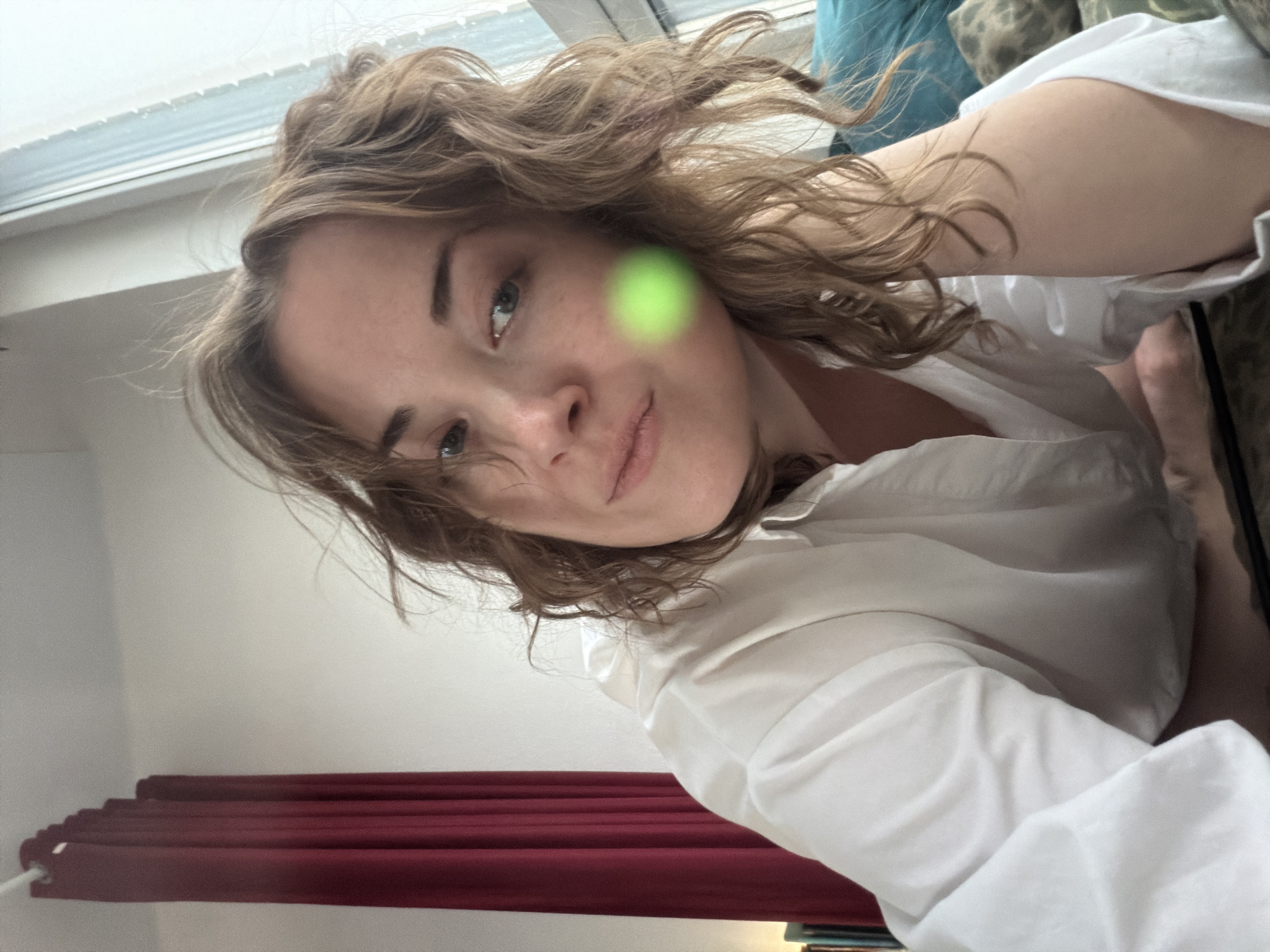 A woman with curly brown hair and fair skin is the primary subject of the image. She is wearing a white button-down shirt and appears to be lying down, possibly on a bed or couch. The camera angle is from above, looking down at her face. The background includes a window with blinds, a wall, and a red curtain. There is a green blur obscuring part of her face. The city and location are unknown.