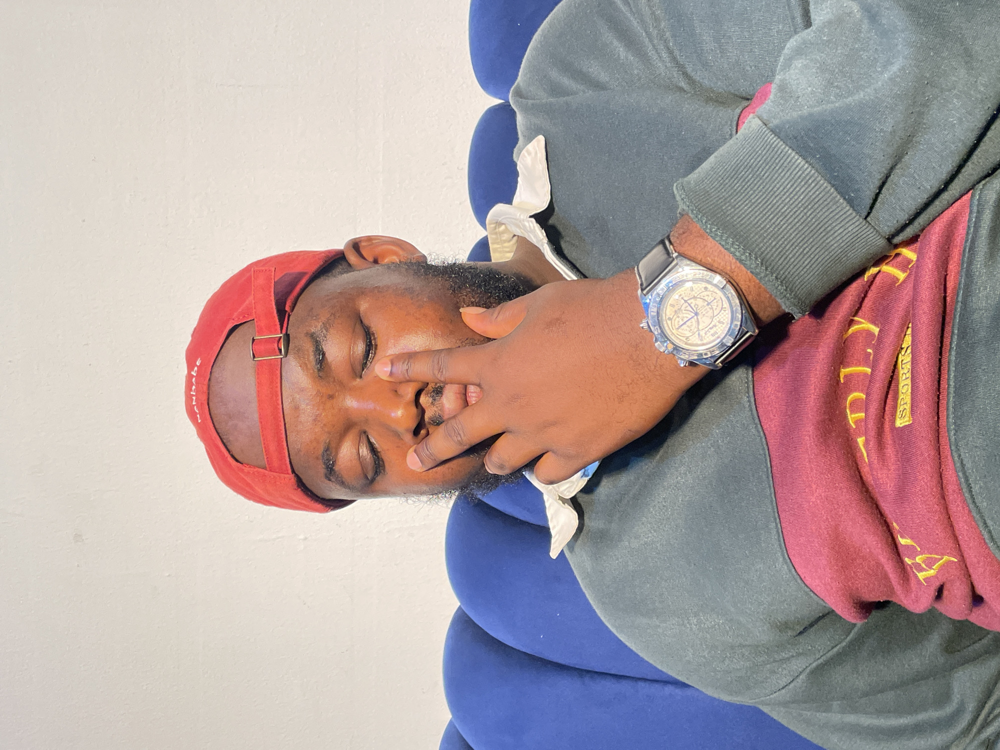A man lies on a blue chair in what seems to be a casual setting. He is wearing a red baseball cap and a sweatshirt with green and maroon sections. His hand is near his face, and he appears to be asleep or resting with his eyes closed. A watch with a black strap is visible on his wrist. The location is Barakin Akawo, Nigeria.