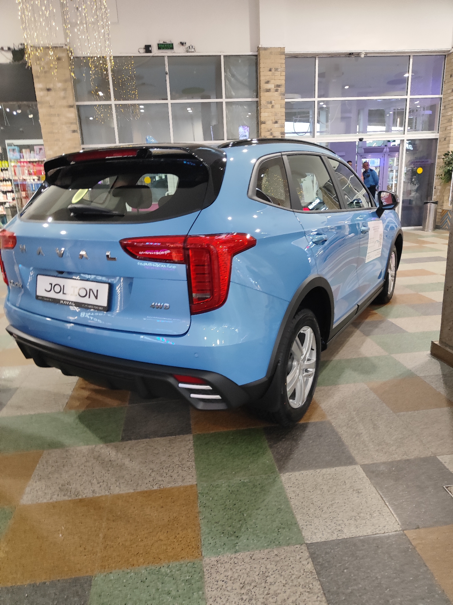 A blue Haval Jolion SUV is parked indoors on a checkered tiled floor. The car is viewed from the rear passenger side. The license plate is visible and reads "JOLION". The car has a black roof and black trim. Behind the car, a glass-fronted building entrance with reflective windows is visible. A person wearing a blue shirt and dark pants is standing in the doorway. Overhead, strings of fairy lights are hanging. The environment appears to be a shopping mall or showroom.
