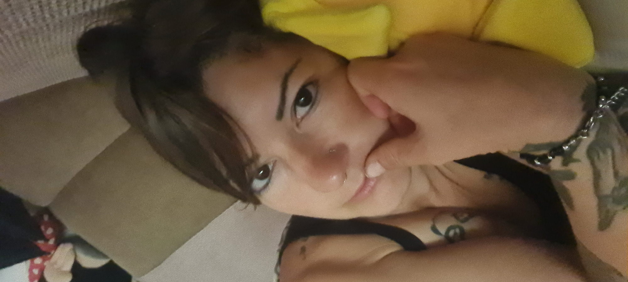 A woman with brown hair and a nose piercing is lying down, resting her face on her hand. She is positioned on a beige pillow or cushion, and a yellow object is visible behind her head. Part of her upper body is exposed, revealing a tattoo on her chest and another tattoo on her arm, along with a chain bracelet. The overall scene appears to be indoors. There is no information in this image to indicate the location is in Nahariyya, Israel.