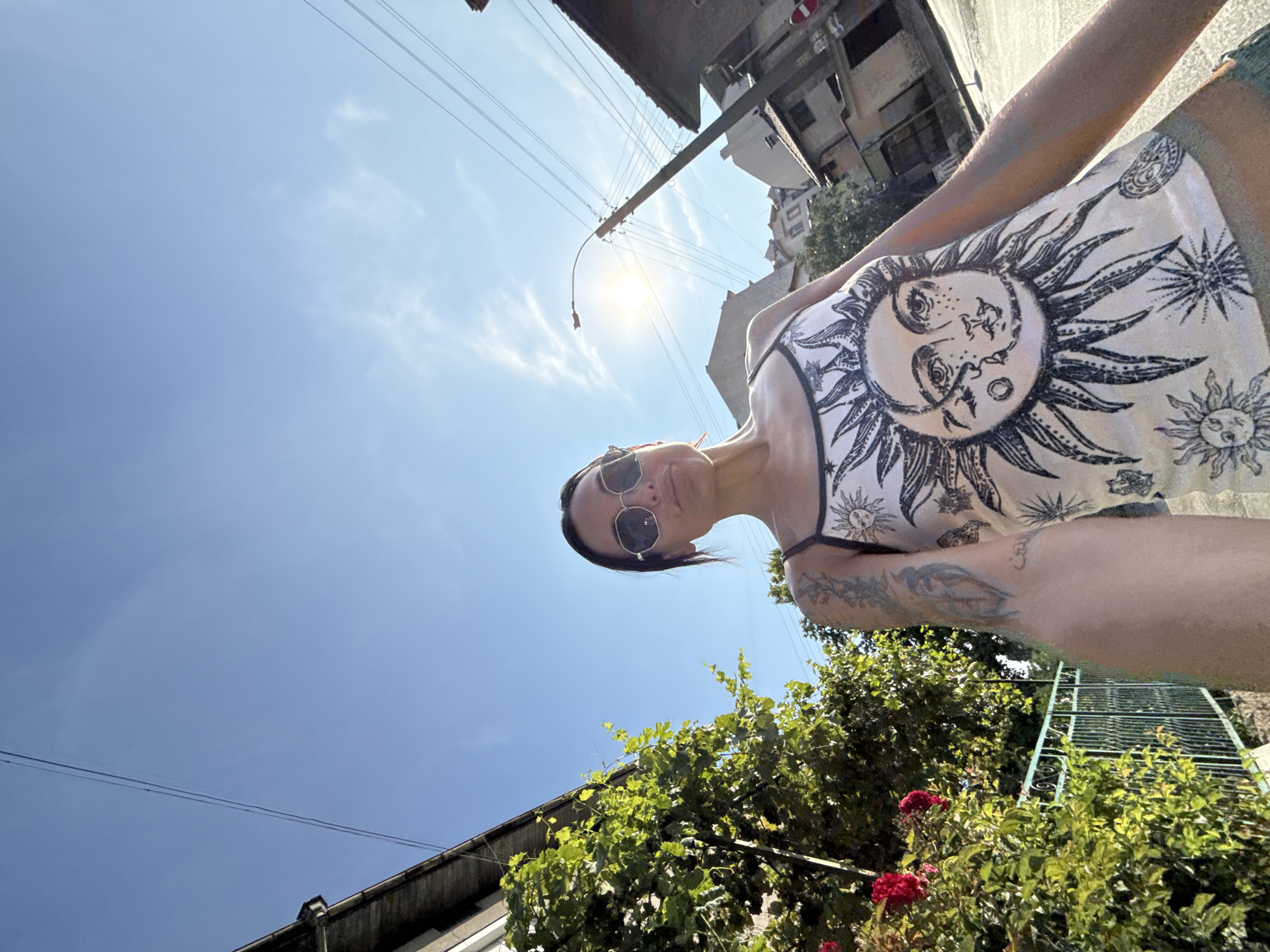 A low-angle, wide shot captures a young woman with dark hair and sunglasses standing on a street in Pleven, Bulgaria, during the day. She is wearing a white tank top with a black sun and moon graphic and blue-green shorts. Her left arm, adorned with tattoos, is extended towards the camera as if taking a selfie. Behind her, a lush green bush with red roses is visible on the left, bordering a light green metal fence. The background features residential buildings with tiled roofs, a utility pole with electrical wires, and a bright sun shining in a clear blue sky with wispy clouds. The street appears to be paved and slightly sloped.