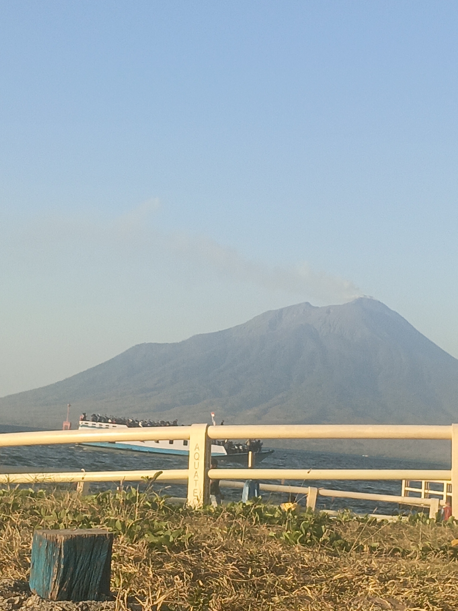This image captures a natural landscape dominated by a large, dormant volcano. A plume of smoke emanates from its summit, suggesting recent volcanic activity. In the foreground, a body of water is visible, with a ferry or large boat carrying numerous passengers. A yellow railing runs horizontally across the frame, with the word "AQUATEX" written vertically on one of its posts. The surrounding vegetation is dry and sparse, with a weathered wooden post in the lower-left corner. The sky is clear and blue. The image is likely taken in or near Lewoleba, Indonesia, given the presence of a volcano and a ferry, common features in coastal communities of the region.