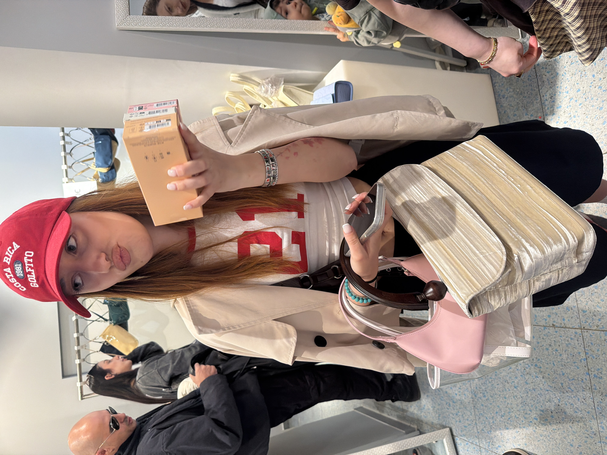 A young woman takes a selfie in what appears to be a clothing store or boutique. She holds a tan box in her left hand and a cell phone in her right hand, with a tan purse with white pleats on her right side. She wears a white shirt with a red number on it, a light brown trench coat, black pants, and a red baseball cap.
In the background, a clothing rack displays various garments. A bald man wearing sunglasses and a black coat stands in the lower left of the frame, and another woman with dark hair stands nearby. The reflections in the mirrors suggest that the store is well-lit. The scene depicts a casual shopping environment. There is no visual evidence in the image that places this scene in Sofia, Bulgaria.