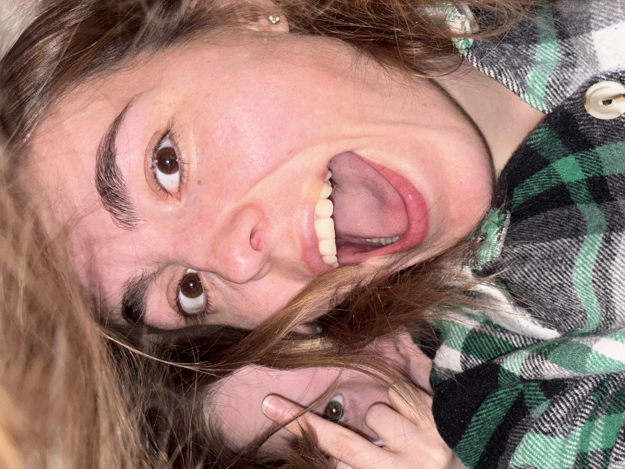 The image shows two individuals in close proximity. One individual's face is visible, turned upside down and with their mouth open wide in what appears to be a playful or expressive gesture. The second individual's face is partially obscured by their hand and hair. The person is covering one of their eyes with their fingers. The individuals are wearing a button-down flannel shirt with a green, white, and black plaid pattern. The surroundings are out of focus and do not appear to include any elements that indicate a specific location, such as the city of Izhevsk, Russia.