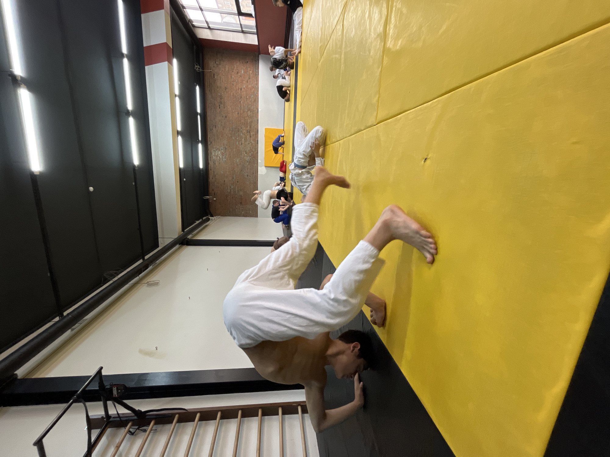 A low-angle shot captures a man in a white karate uniform, upside down against a yellow wall, seemingly performing a handstand or acrobatic maneuver. His face is obscured, and his body is angled, with his bare feet in the upper part of the frame.
In the background, slightly out of focus, are other people in similar uniforms, some standing and others lying on what appears to be a padded floor, potentially practicing martial arts or acrobatics.
The location appears to be an indoor training facility with a brick wall visible, windows towards the top, and a wall with strip lighting. The picture's unique angle and perspective make it challenging to pinpoint an exact location within Krasnoyarsk, Russia, without additional context.