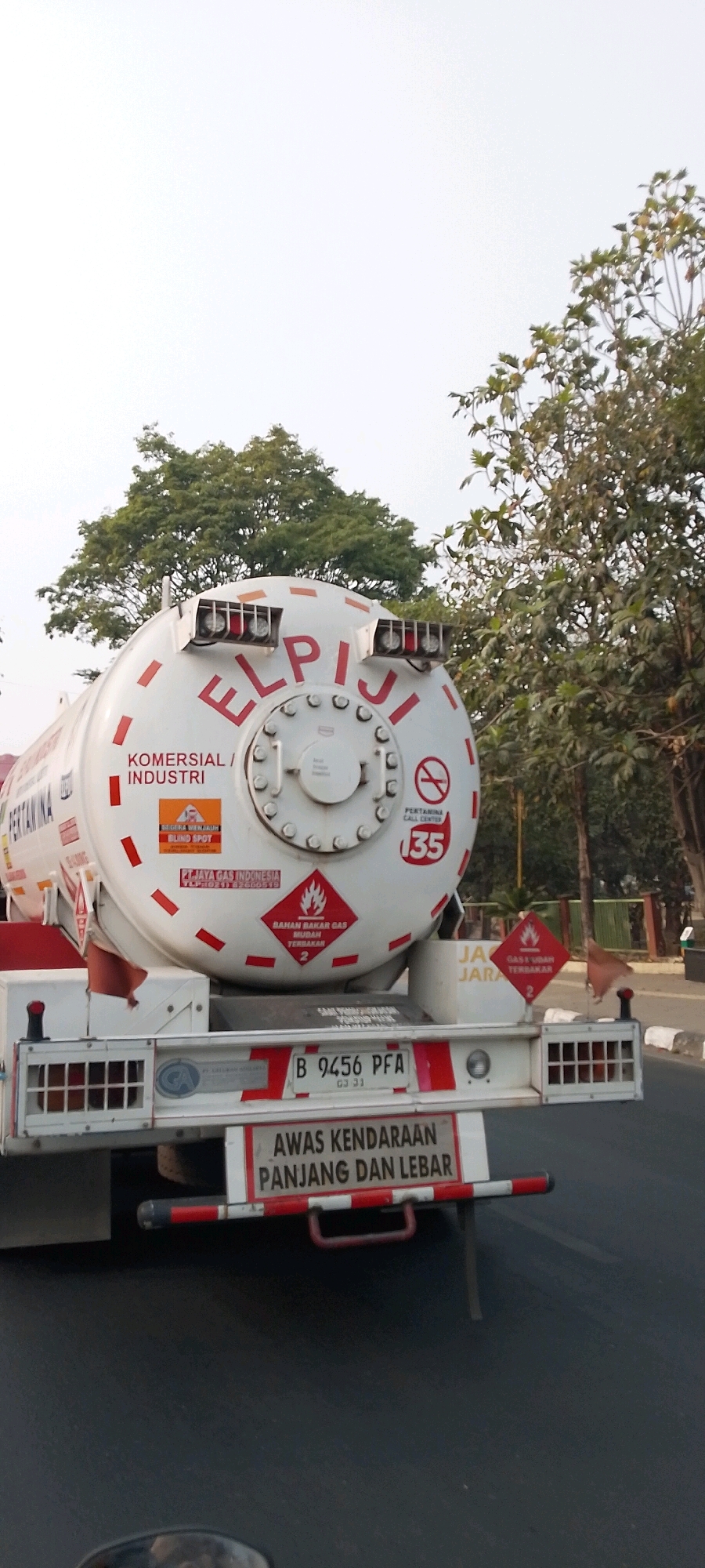 The image shows a Pertamina branded tanker truck, likely carrying liquefied petroleum gas (LPG), in Bekasi, Indonesia. The truck has prominent "ELPIJI" lettering on its tank, which is a common brand name for LPG in Indonesia. Various safety labels are visible, including hazard symbols for flammable gas and warnings for "LONG AND WIDE VEHICLES". A license plate that reads "B 9456 PFA" is also visible. The truck appears to be on a road with trees and some buildings in the background, suggesting an urban or suburban environment within Bekasi. The sky is overcast.