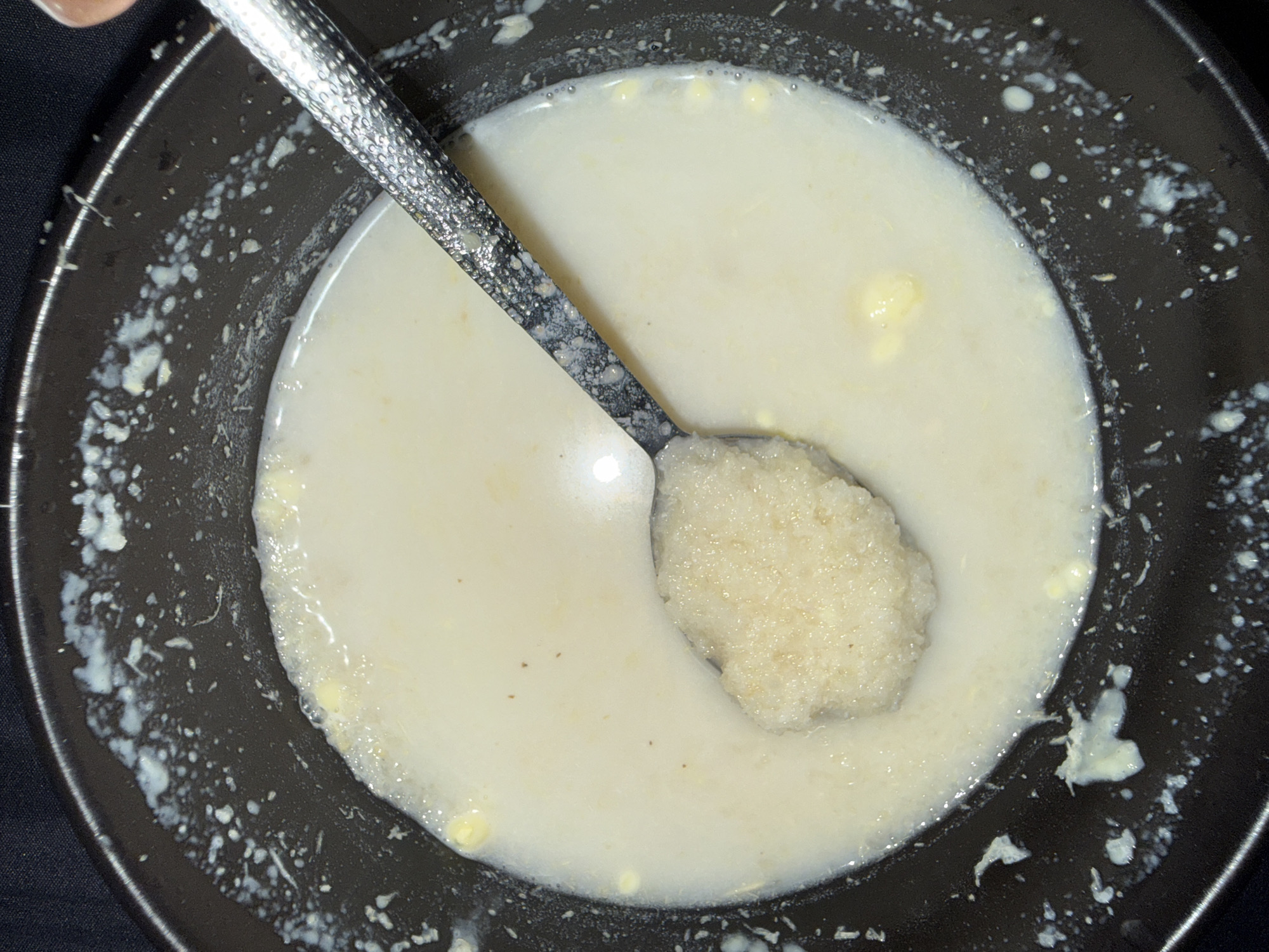 An overhead, close-up shot shows a dark bowl filled with a light-colored, creamy substance, likely oatmeal or porridge. A spoon, with a textured metallic handle, is partially submerged in the bowl, scooping up a mound of the same creamy substance. Small yellow flecks, possibly butter or corn kernels, are visible throughout the porridge. The rim of the bowl has white splatters, suggesting it may have been recently filled or stirred vigorously. The background is a dark, soft surface, possibly fabric. There are no visible indications of the city of Abuja, Nigeria, or any other specific geographical location.