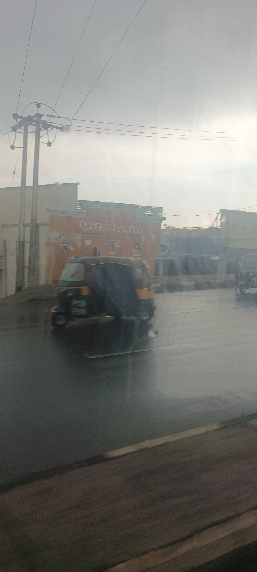 The photograph appears to be taken from inside a moving vehicle, possibly a car or bus, looking out towards a street scene. The location is indicated by the presence of an "Vitafoam" advertisement on a building, which is a company known to operate in Nigeria, including Jos.
The visible street is wet, suggesting it has recently rained. There are at least two "Keke Napep" tricycle taxis on the road, one of which is partially covered. Power lines are visible overhead, supported by a utility pole on the left side of the frame. In the background, there are other buildings, some of which are blurred, giving a sense of depth and movement.
The overall impression is one of a wet, somewhat overcast day in an urban environment in Jos, Nigeria.