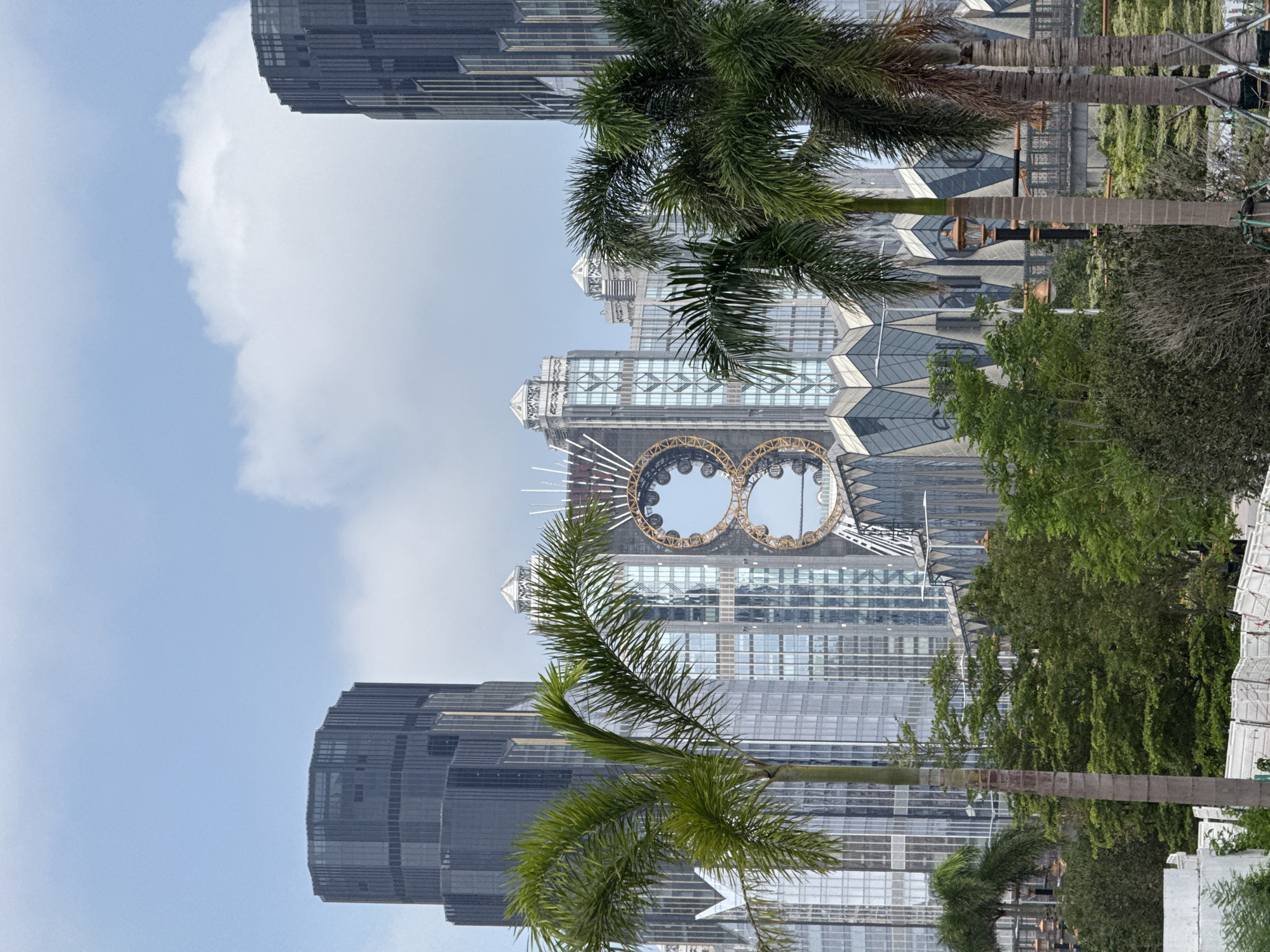 A daytime medium shot captures a cityscape in Unknown, Macao, featuring the iconic Studio City complex. The architectural highlight is the figure-eight-shaped Ferris wheel suspended between two towers. Adjacent to this structure are taller, dark-colored skyscrapers. Lush palm trees frame the scene, with their fronds partially obscuring the buildings. The sky is partly cloudy, with large white clouds contrasting against the light blue. The overall environment suggests a modern urban area with tropical influences.
