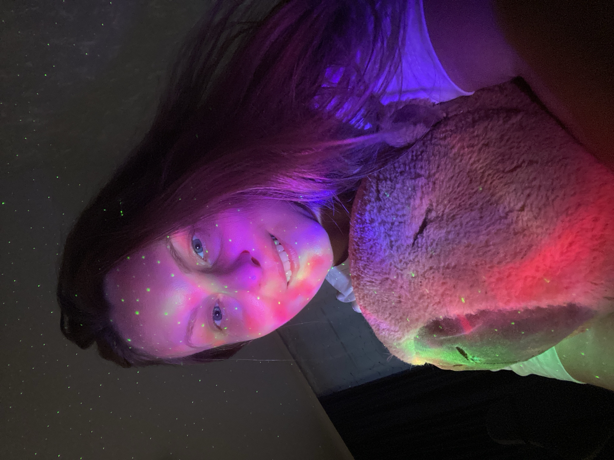 In a dimly lit room, a young woman with long, dark hair is illuminated by colorful lights and small green specks that resemble stars. She is holding a fluffy, pink stuffed animal close to her chest. Her face is turned towards the camera, and she is smiling. The lighting creates a dreamlike atmosphere, with hues of pink, purple, and green washing over her face and the stuffed animal. The background is dark, with hints of what might be a window or wall visible on the left side of the image. This scene appears to be taking place indoors, possibly in a bedroom, and there are no indications of the specific city of Noviy, Russia.