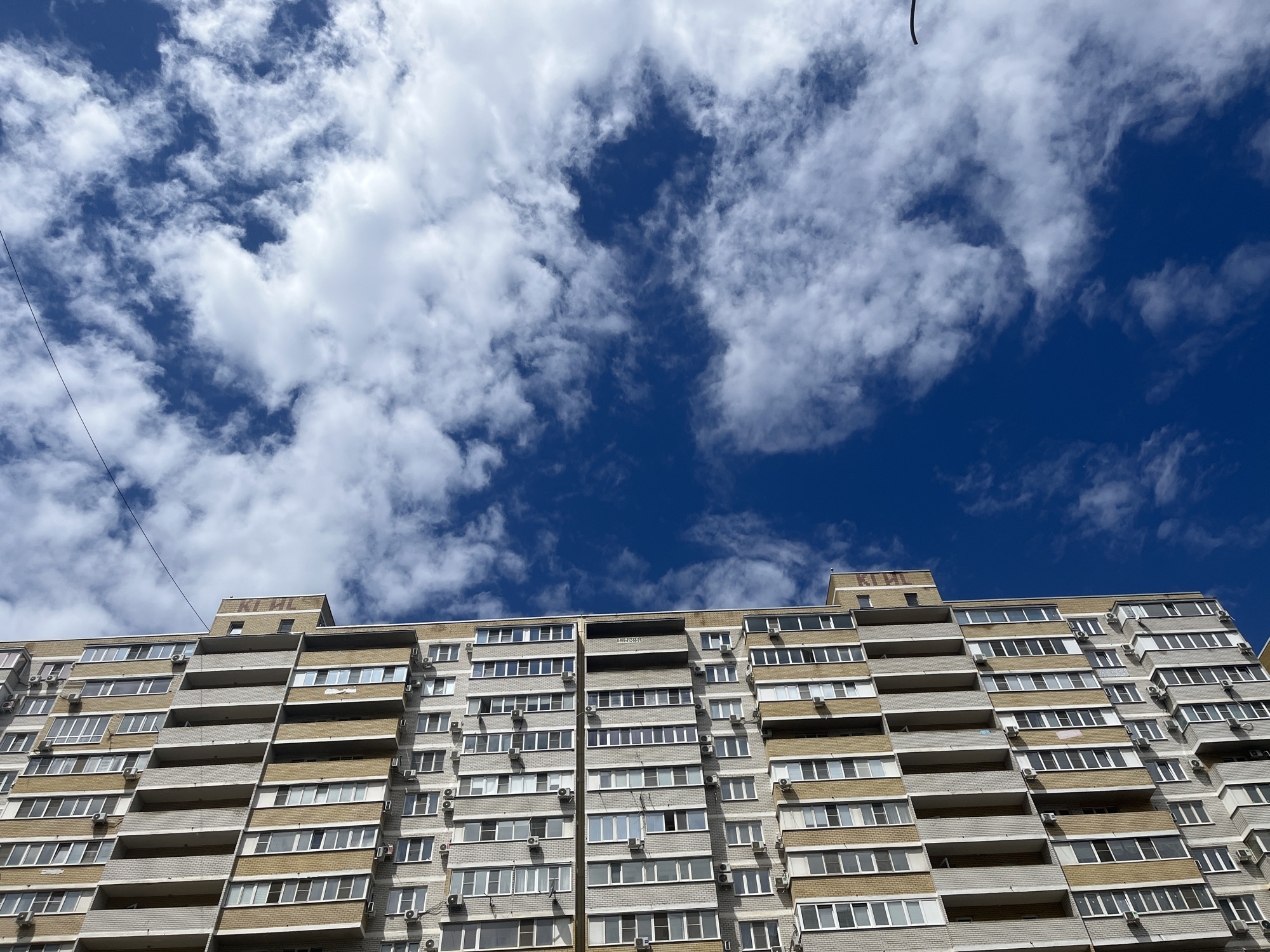 The image shows a tall apartment building in Krasnodar, Russia, under a blue sky with scattered white clouds. The building has multiple floors with numerous windows and balconies, and is constructed of gray and light brown brick. Many of the units have air conditioning units mounted on the exterior walls. A power line is visible diagonally across the upper left portion of the image. The name of the building (КГПИ) is inscribed on the top of the building.