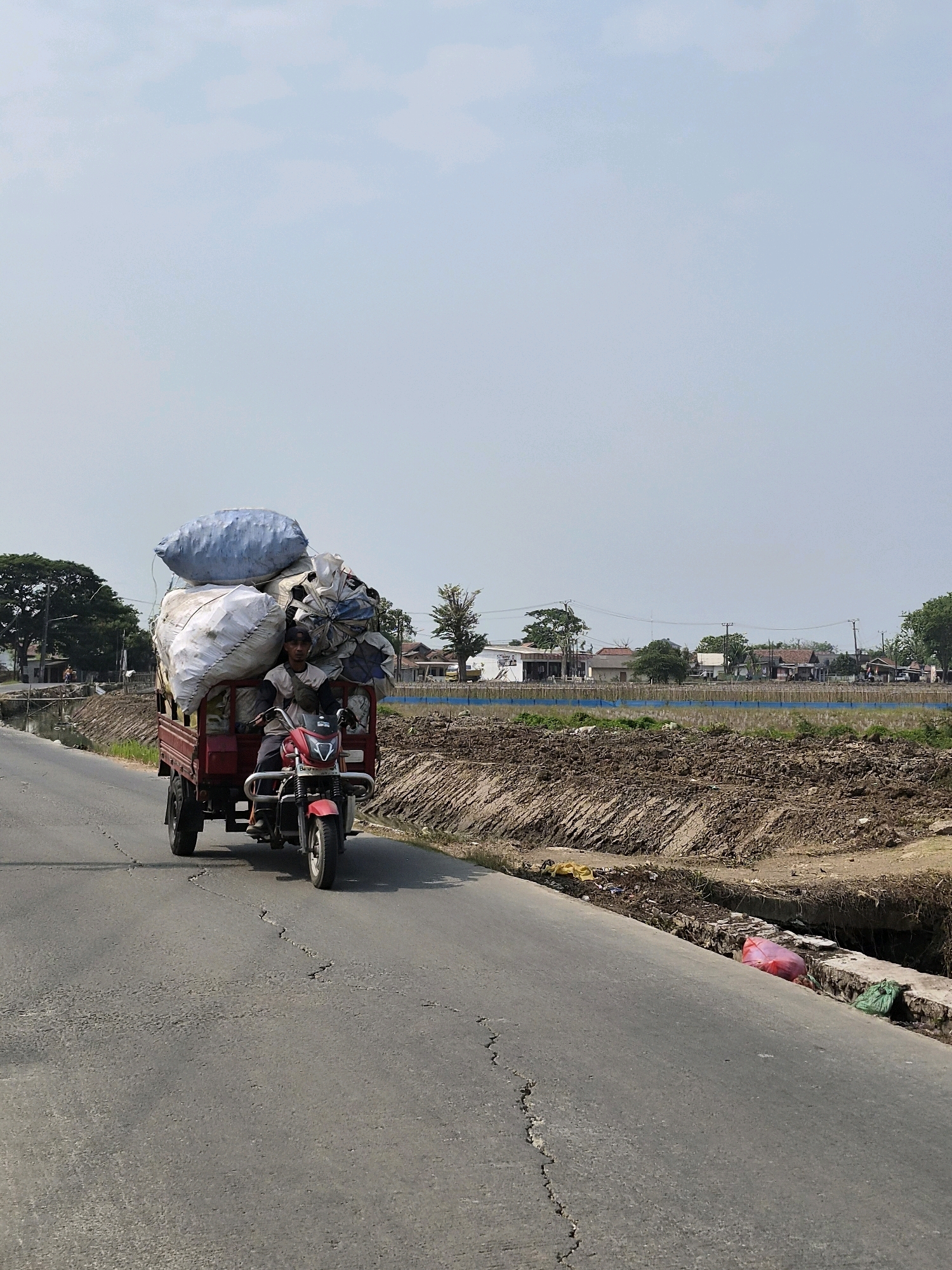 In Srimahi, Indonesia, a man is riding a red three-wheeled motorcycle loaded with large sacks of material. The motorcycle is traveling on an asphalt road with some cracks. To the right of the road is a ditch filled with debris, including plastic bags. Beyond the ditch is a muddy field with some vegetation and a few buildings in the distance. To the left of the road, there is a body of water and trees. The sky is clear and blue.
