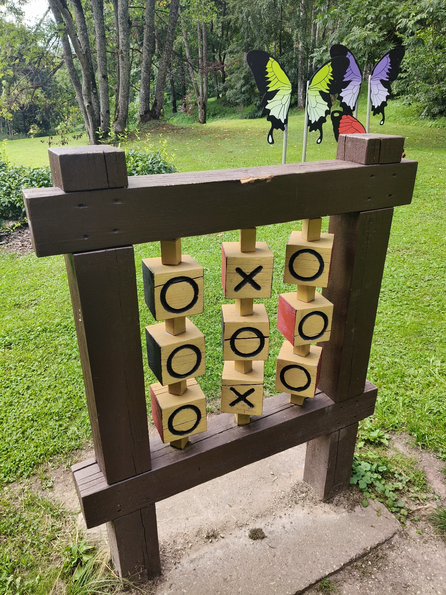A large wooden tic-tac-toe game is set up outdoors in a park-like setting. The game features a brown wooden frame supporting nine wooden cubes, each rotatable to display either an 'X' or an 'O'. The cubes are yellow with black markings, and some also have red sides. The game is situated on a concrete pad surrounded by green grass and trees. In the background, there are decorative cutouts of large butterfly wings in vibrant colors. This scene is located at the Koknese Open-Air Stage and Park in Koknese, Latvia. There are no people visible in the image.