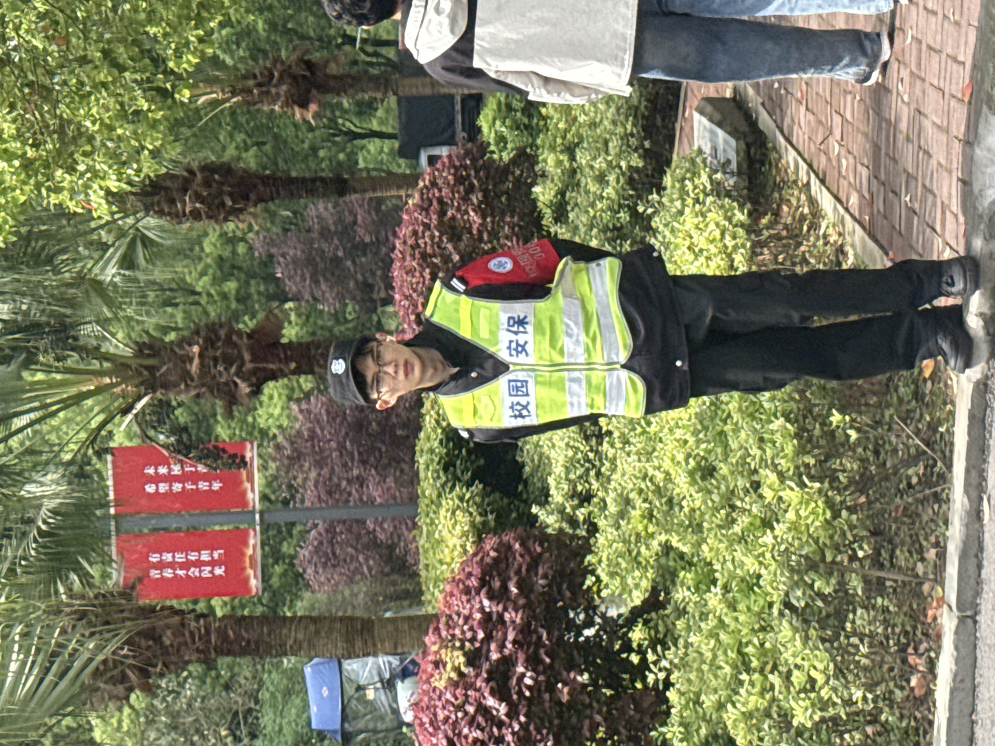 The image shows a person wearing a security guard uniform with "校园安保" (campus security) written on the vest, standing outdoors with various trees and bushes in the background. A red sign is visible to the left with Chinese characters that include the phrase "青春才会闪光" (youth will shine). The location is Jiangjin, China. Other people are visible in the background.