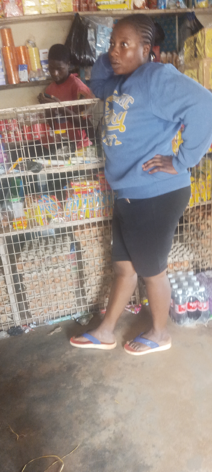 A woman wearing a blue sweatshirt and black shorts stands with her left arm resting on a wire cage in a small store in Bokkos, Nigeria. She is looking towards the right. Behind the cage are assorted goods including snacks and eggs. A child in a red shirt stands behind her, and a shelf of merchandise is visible above them. Plastic bottles of soda are on the floor. The ground is a worn, undecorated concrete.